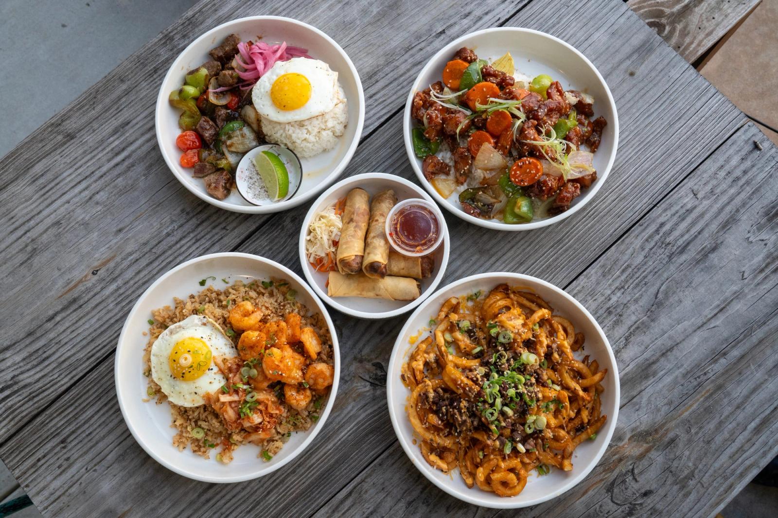 Five bowls on a wooden table, including a beef rice bowl with egg, shrimp fried rice, a beef and vegetable dish, spring rolls with dipping sauce and stir-fried noodles.