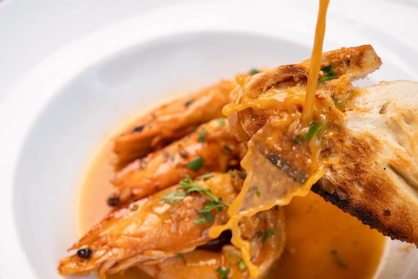 Sauce is poured over grilled head-on prawns and toasted bread in a white bowl garnished with herbs.