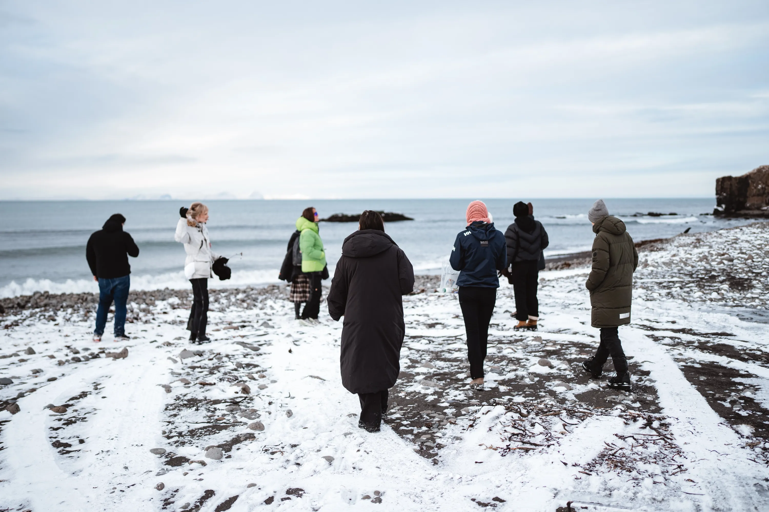 Un gruppo di persone cammina su una spiaggia di sassi scuri parzialmente coperta di neve, dirigendosi verso la riva del mare. Indossano abbigliamento invernale pesante, inclusi parka colorati e berretti. Sullo sfondo, il mare calmo sotto un cielo nuvoloso e, in lontananza, il profilo di montagne innevate.
