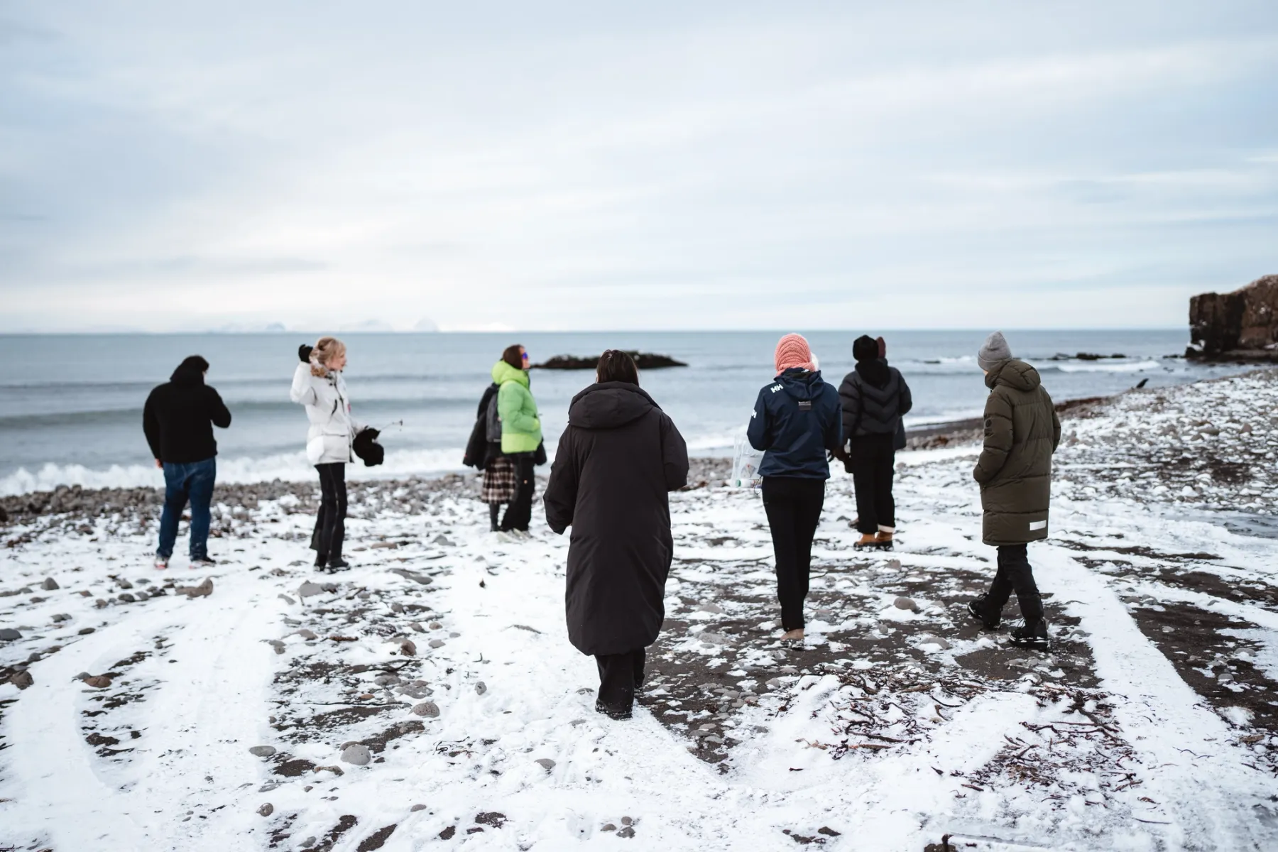 Un gruppo di persone cammina su una spiaggia di sassi scuri parzialmente coperta di neve, dirigendosi verso la riva del mare. Indossano abbigliamento invernale pesante, inclusi parka colorati e berretti. Sullo sfondo, il mare calmo sotto un cielo nuvoloso e, in lontananza, il profilo di montagne innevate.
