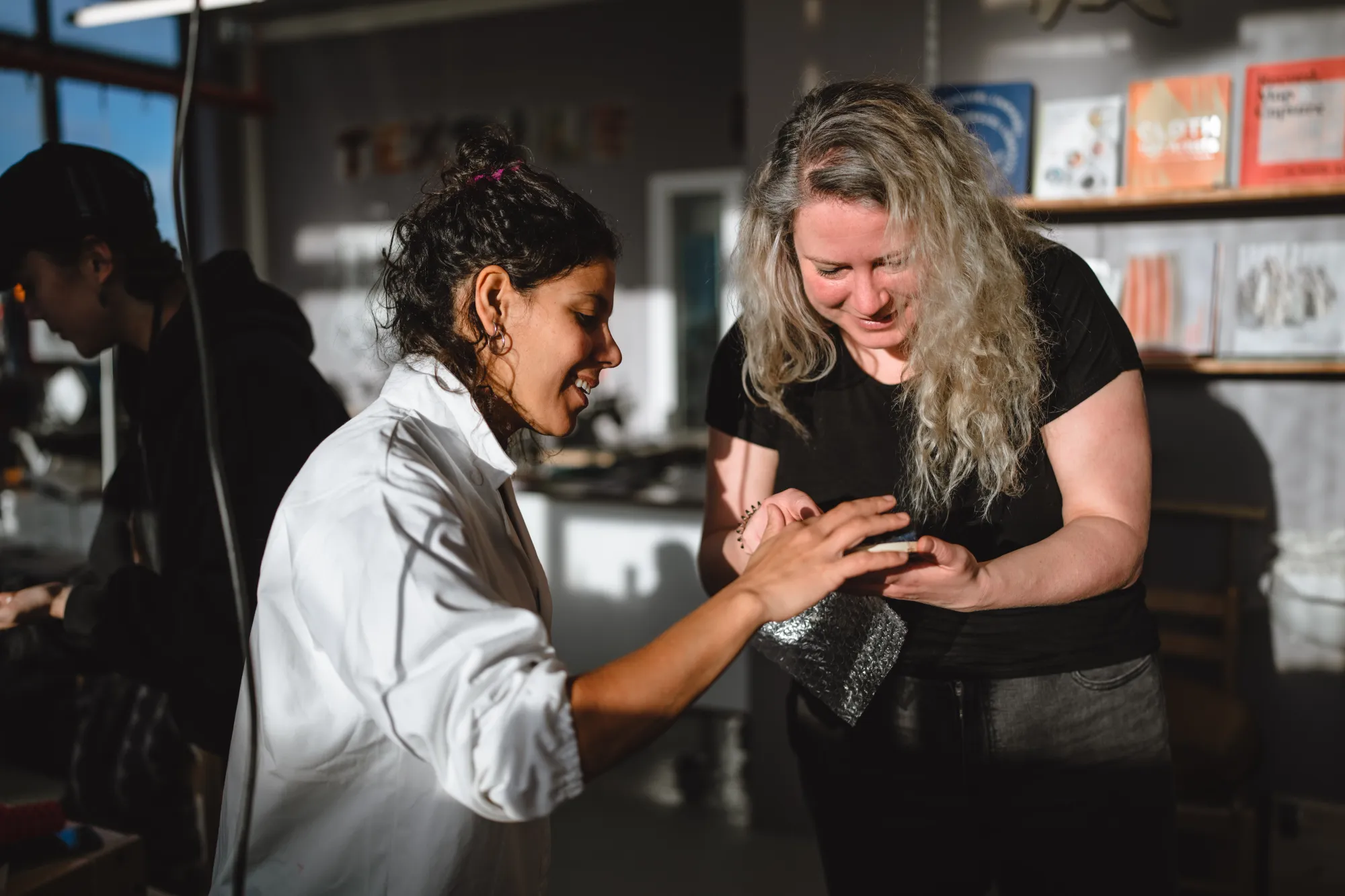 Due donne esaminano insieme un piccolo oggetto o un campione di materiale in un laboratorio ben illuminato. Una donna indossa un camice bianco da lavoro, mentre l'altra una maglietta nera; entrambe sono concentrate sull'oggetto tenuto tra le mani. Sullo sfondo si intravede un ambiente creativo con scaffali pieni di libri e materiali, oltre a una terza persona parzialmente visibile che lavora in ombra.