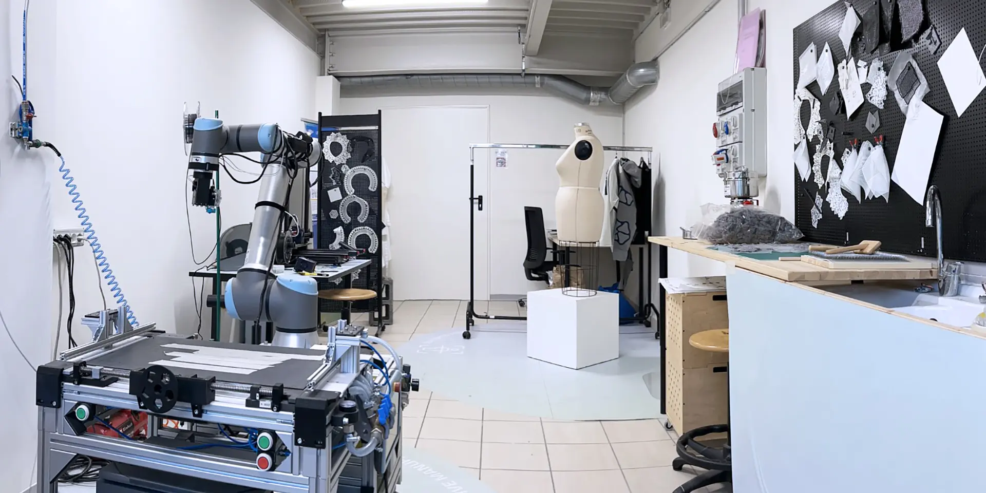 Interior of a fashion tech lab featuring an industrial robotic arm, a 3D printer, a dress form, and a pegboard displaying fabric samples and laser-cut prototypes.