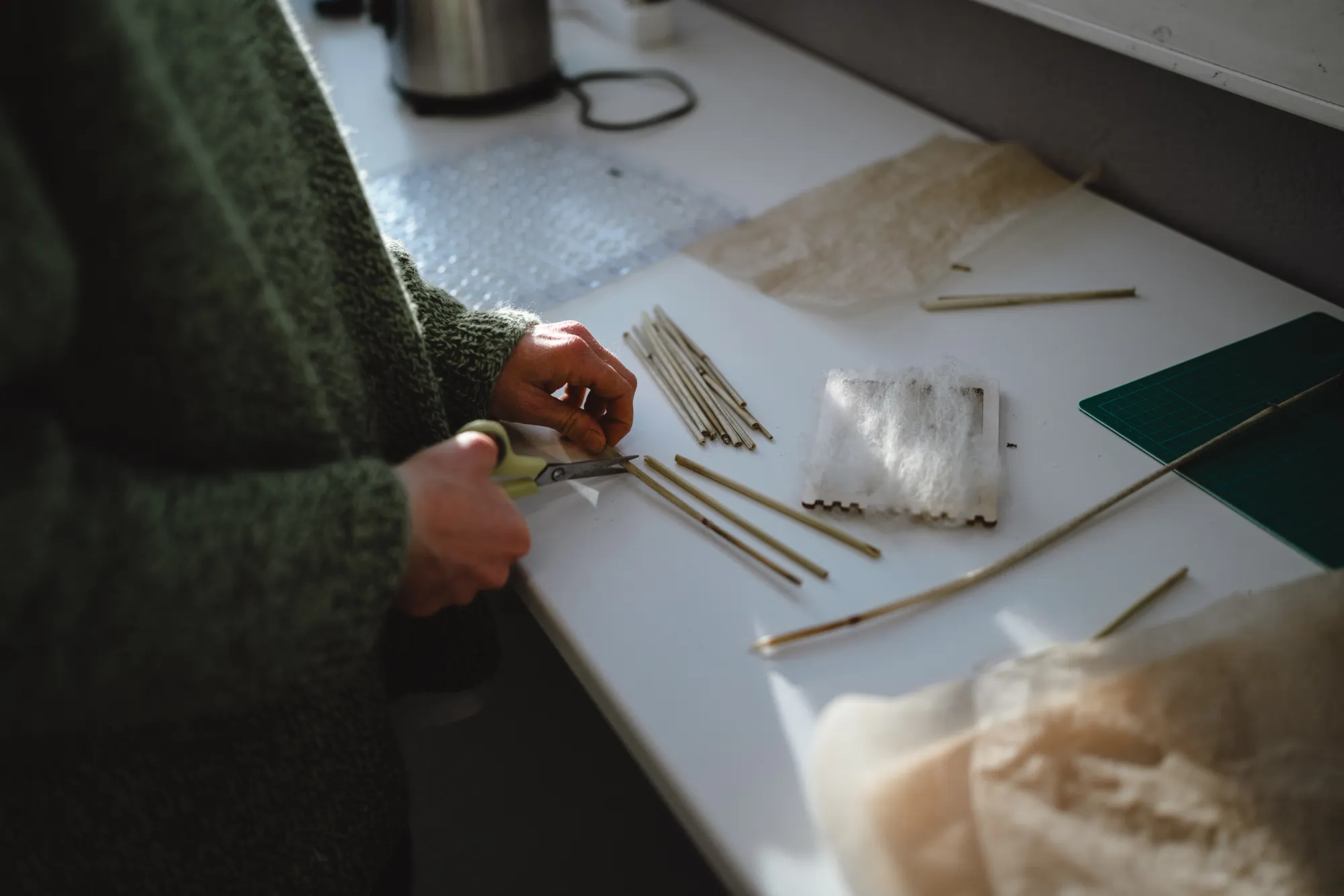 Inquadratura ravvicinata di una persona che indossa un maglione di lana verde mentre lavora a un tavolo. Con un paio di forbici, sta tagliando con precisione dei sottili steli vegetali o rametti. Sul piano di lavoro bianco sono sparsi diversi materiali: piccoli fasci di legnetti, fibre tessili bianche soffici simili a lana, ritagli di tessuto leggero e un tappetino da taglio verde.