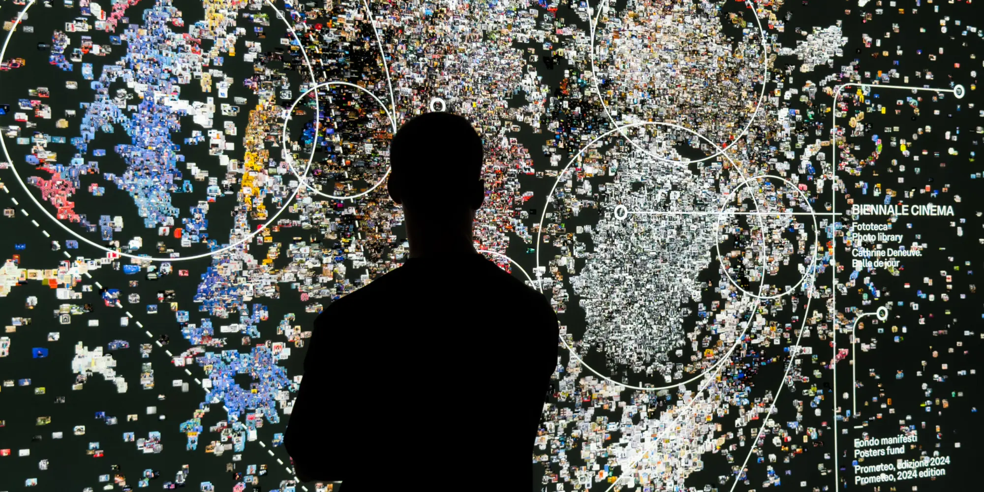 Silhouette di una persona davanti a una grande installazione digitale composta da migliaia di immagini e frammenti visivi colorati, disposti come una mappa astratta su sfondo scuro con linee circolari sovrapposte.