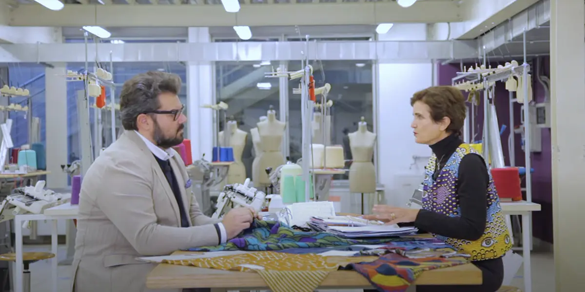 The image portrays a man and a woman engaged in a professional consultation within a modern fashion studio. Seated across from each other at a large wooden table, they are carefully analyzing various brightly printed fabric swatches and technical catalogs. The background, characterized by industrial lighting, clearly reveals the nature of the setting through the presence of dress forms, spools of colored thread, and sewing machinery, suggesting a crucial phase of creative design or material selection for a collection.