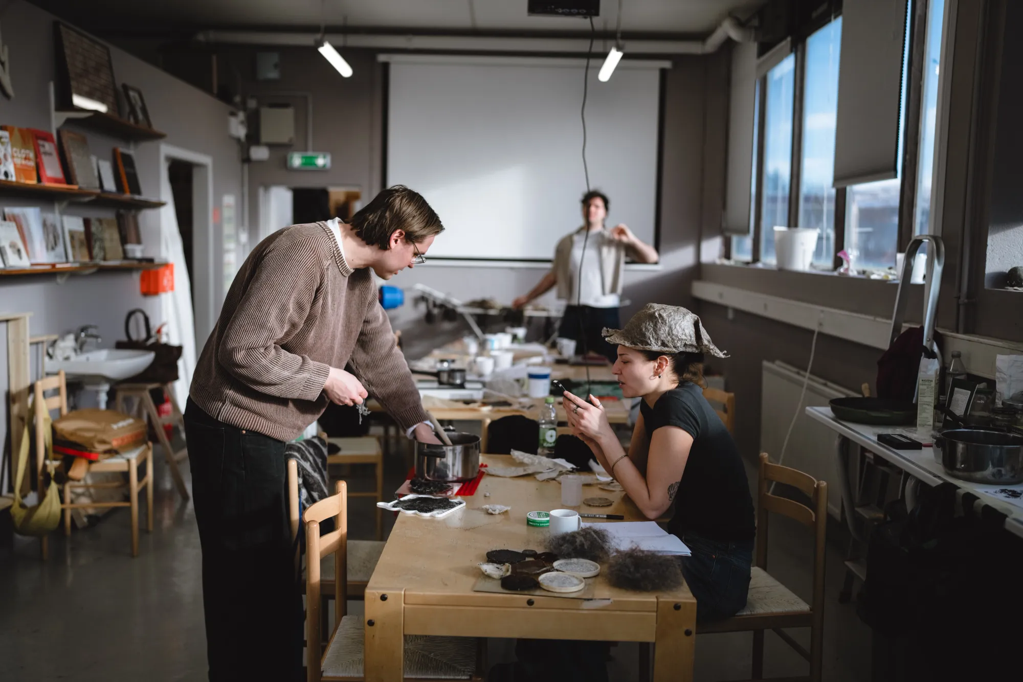 Studenti al lavoro in un laboratorio di design; una ragazza con un cappello sperimentale in biomateriale consulta lo smartphone mentre un collega mescola dei composti in una pentola. Sul tavolo sono presenti campioni di fibre e materiali in fase di sviluppo.