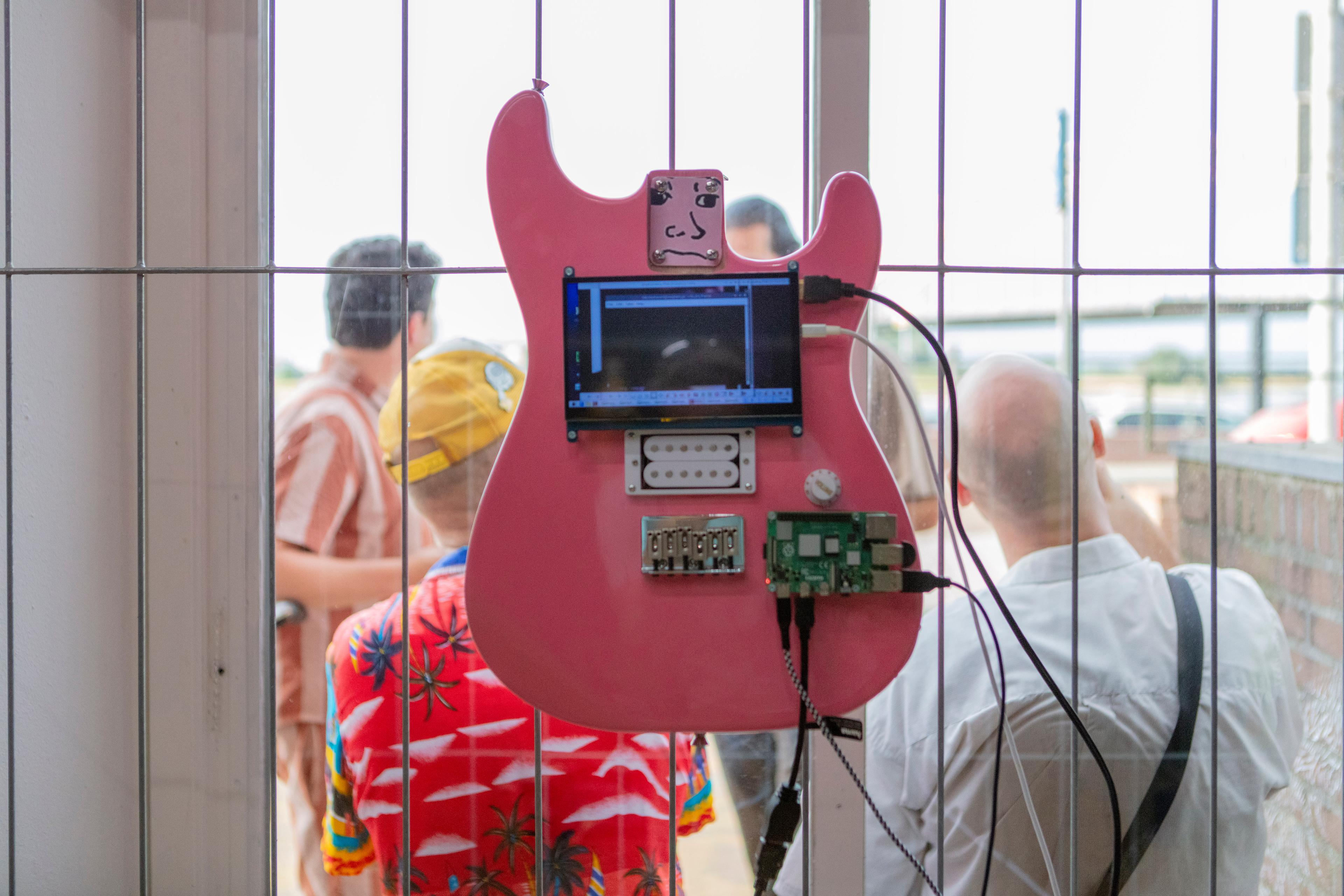 Een roze elektrische gitaar hangt aan een ijzeren hek voor een raam. Aan de andere kant van het raam zien we publiek met hun rug naar de fotograaf staan.