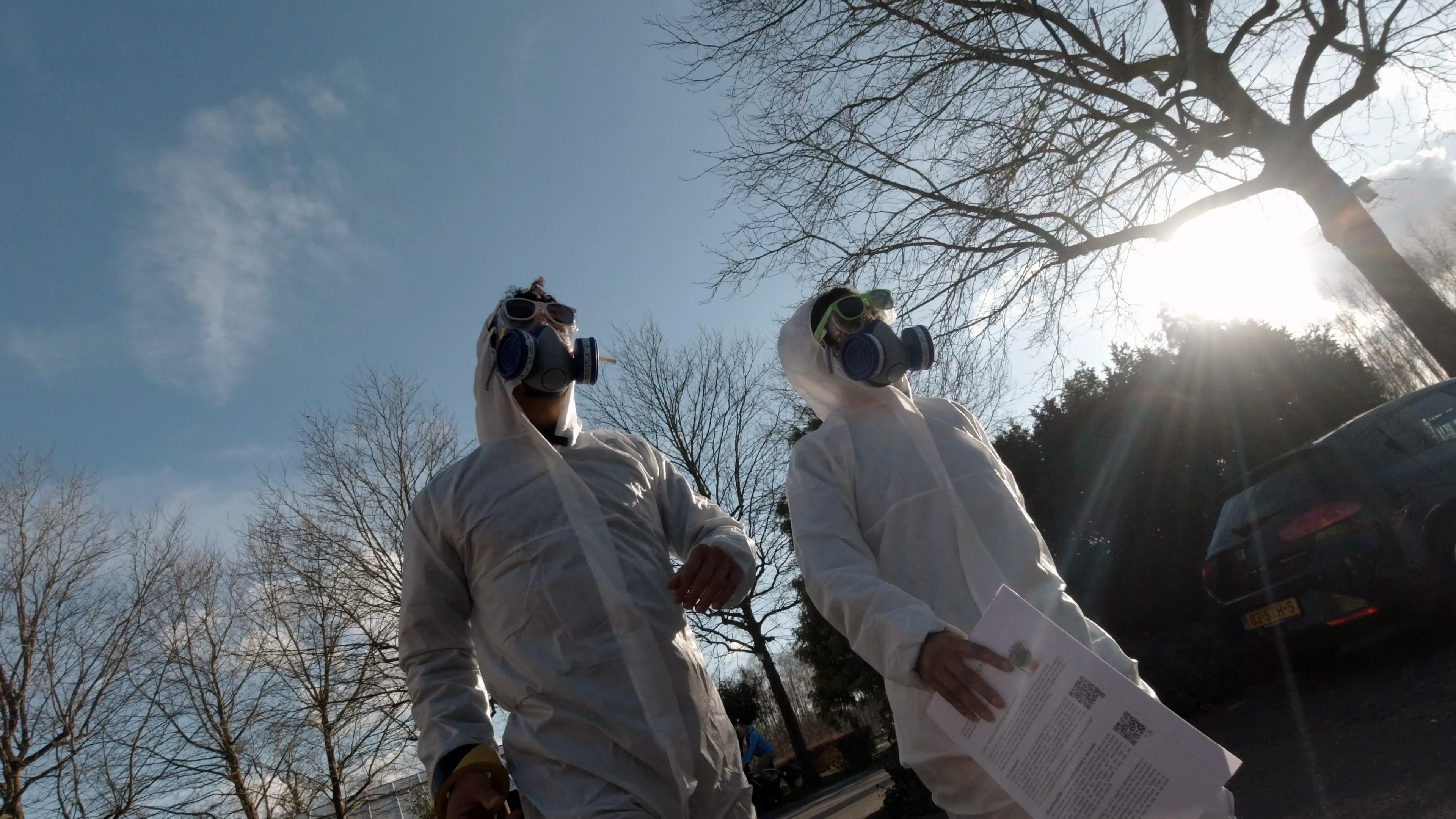 Beelden uit de Youtube serie, de presentatoren lopen in witte pakken met een gasmasker op straat. 