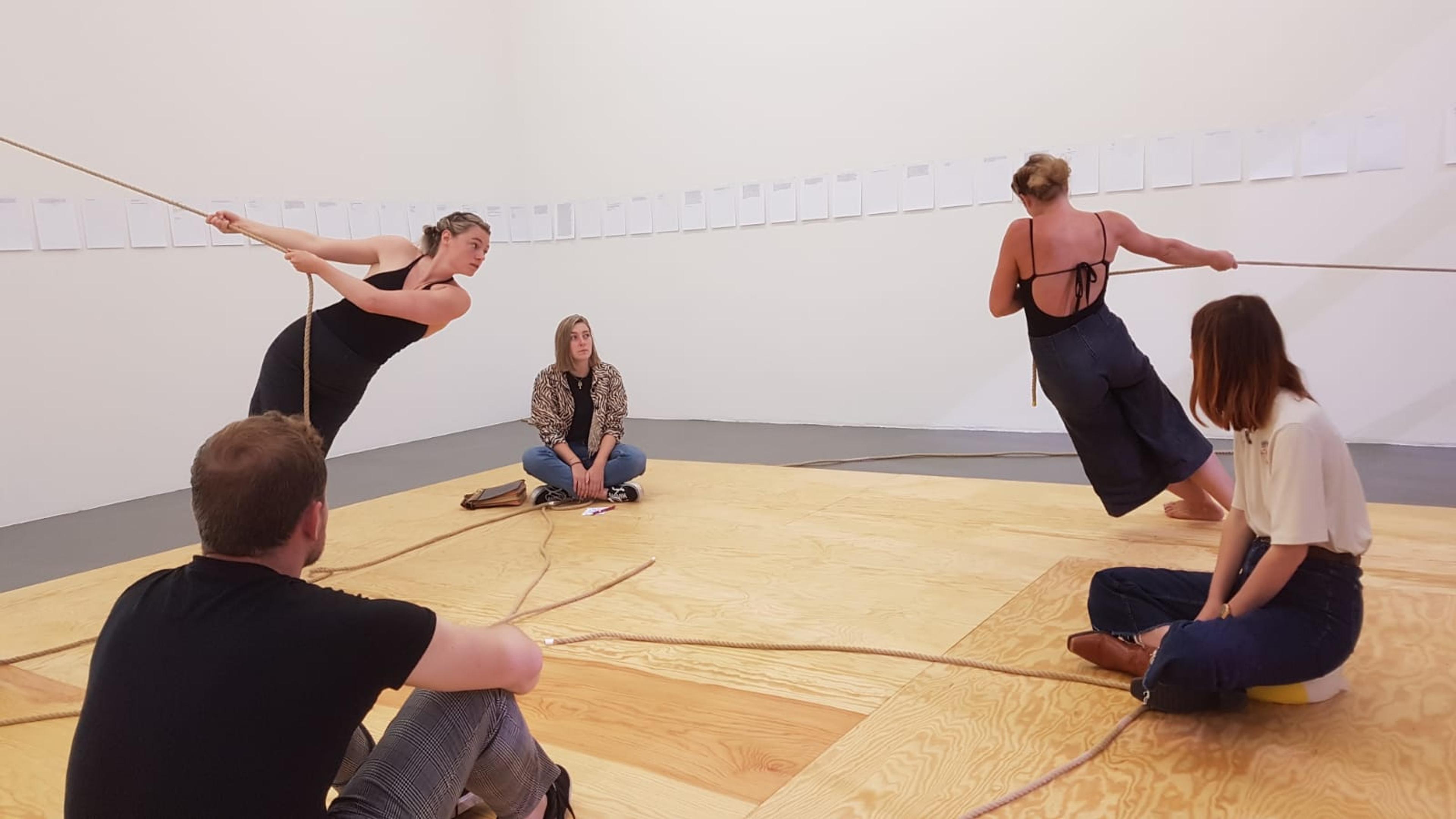 Beeld van de voorstelling LEUN, toeschouwers kijken naar 2 performers die beide een eigen stuk touw vast hebben en daar aan hangen