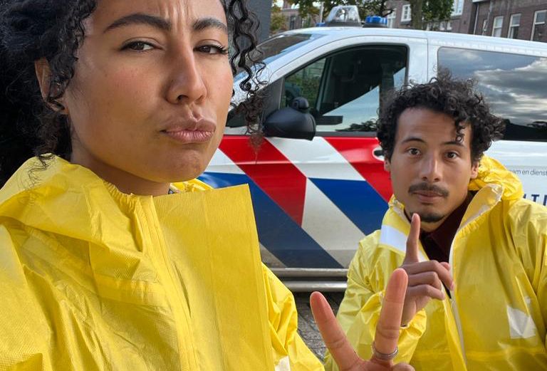 Beelden uit de YouTube serie, de presentatoren staan in gele pakken voor een politiebusje.