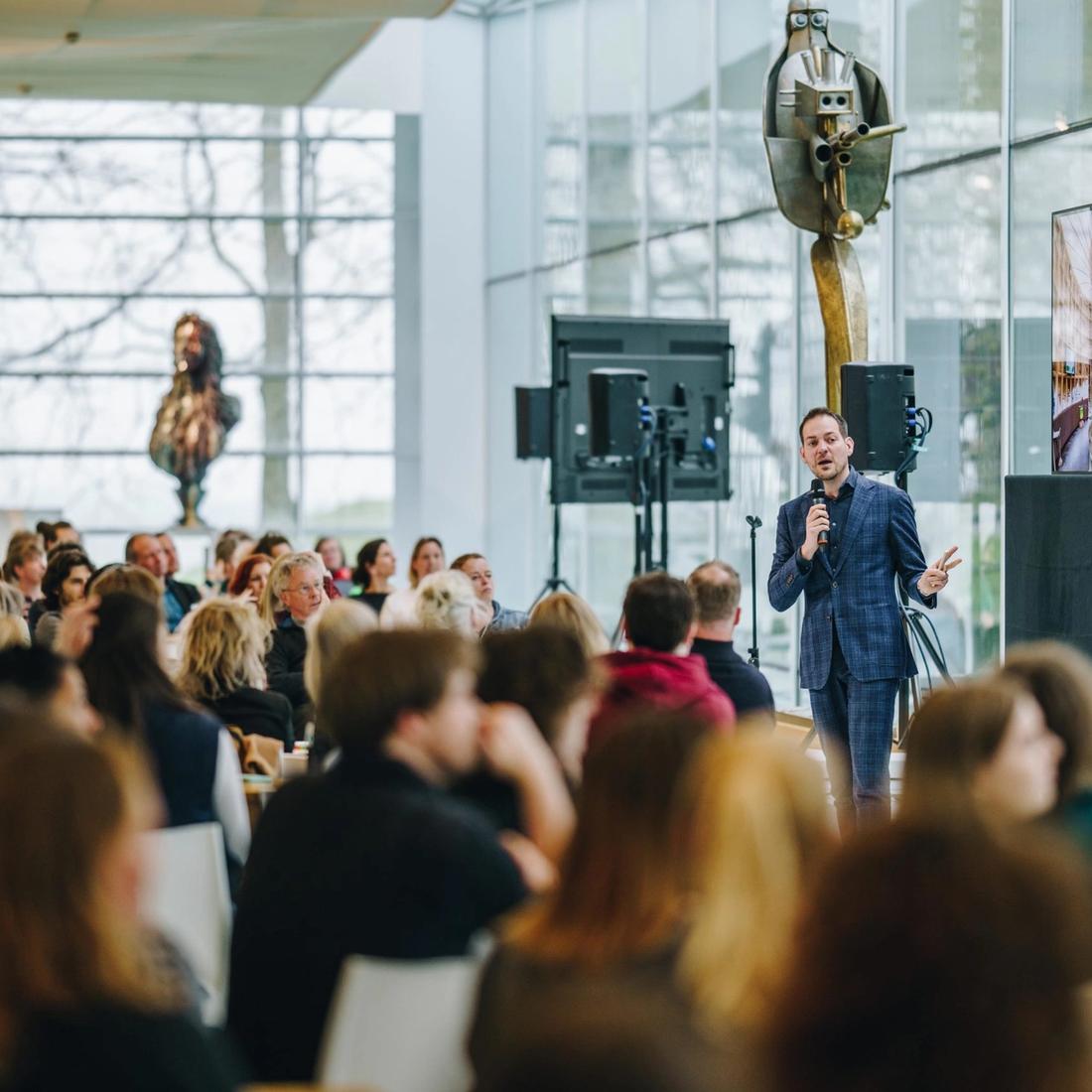 Foto van Lucas de Man, sprekend op een podium voor een zal vol mensen