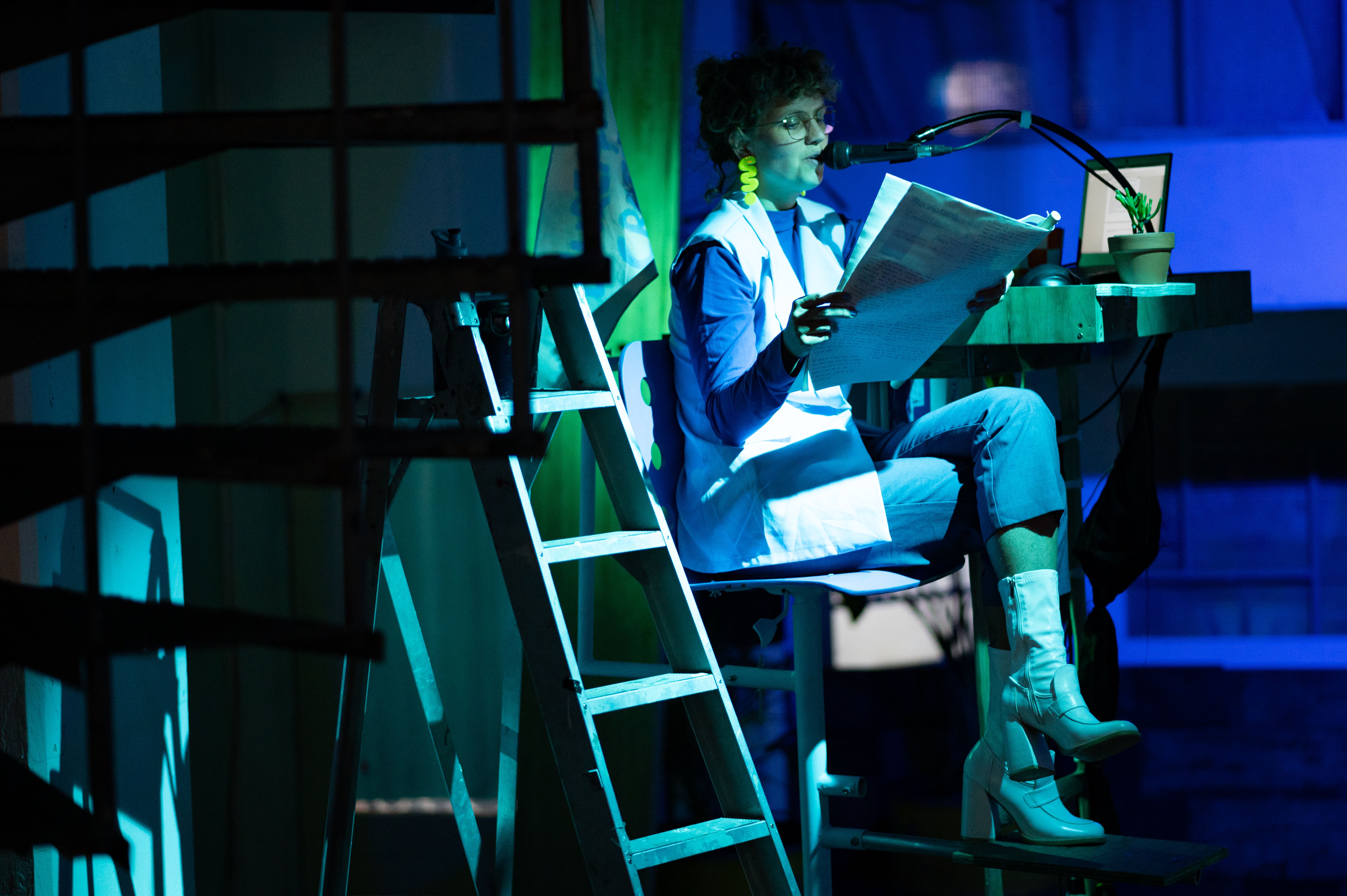Here is a scene from the 2024 production of MOSs Social Club. A woman is sitting on a chair on stage, her legs crossed, reading the newspaper. The lighting is set to shades of blue and green, giving the scene a magical atmosphere. 