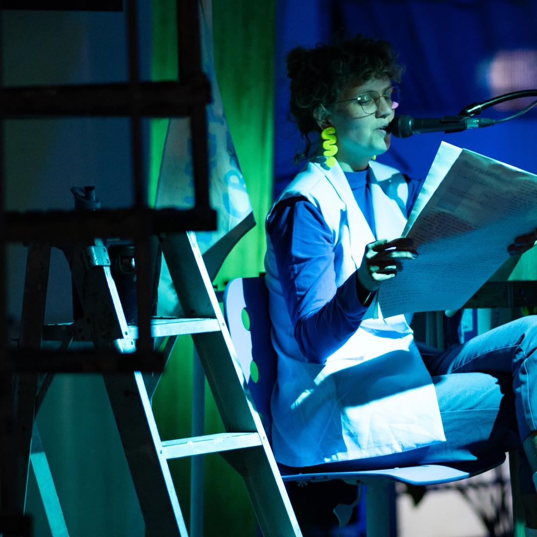 Here is a scene from the 2024 production of MOSs Social Club. A woman is sitting on a chair on stage, her legs crossed, reading the newspaper. The lighting is set to shades of blue and green, giving the scene a magical atmosphere.