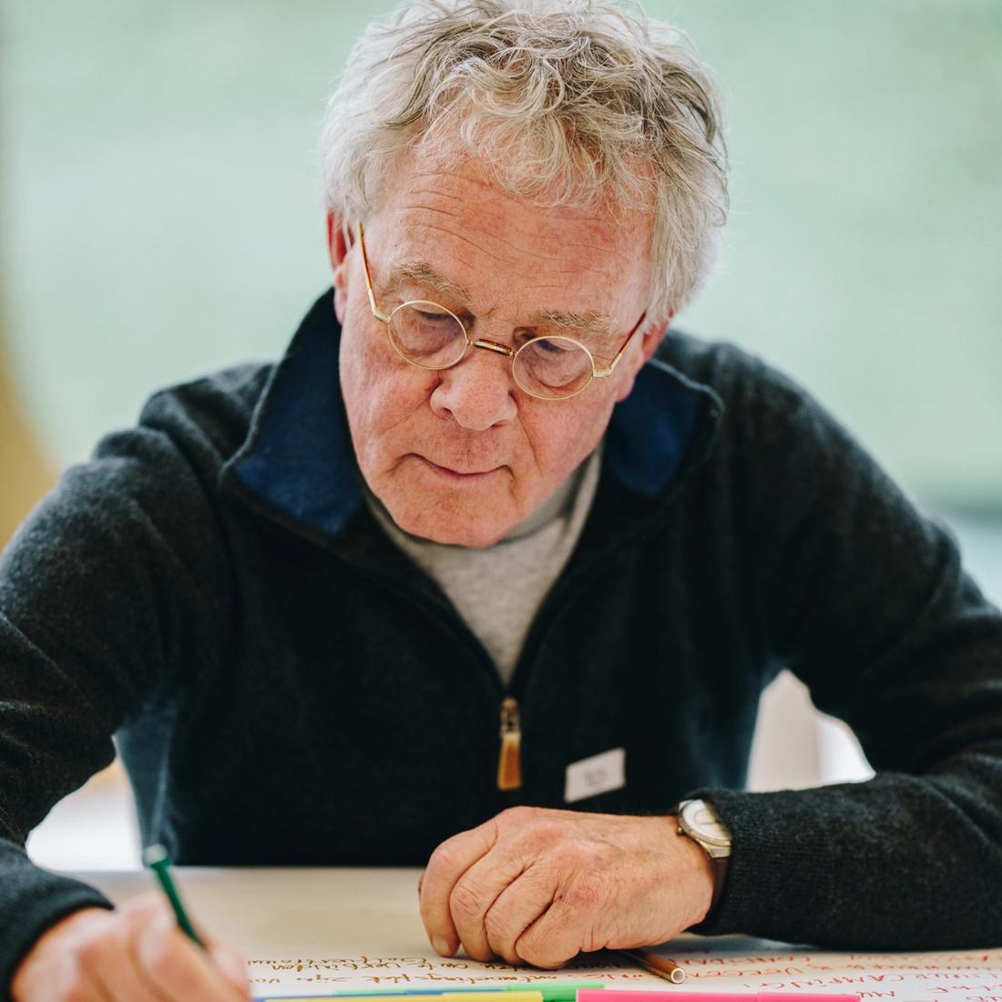 Oudere man met bril die een plan maakt op een groot vel papier met verschillende kleuren stift.