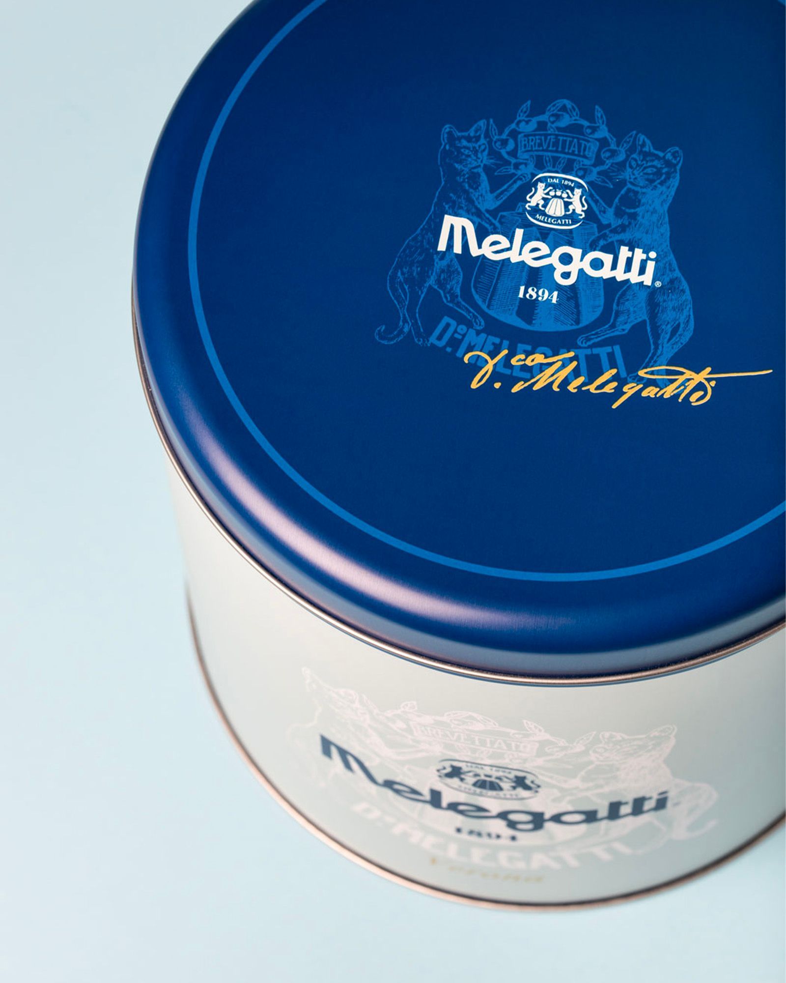 White metal tin with blue lid, decorated with the Melegatti crest logo and signature, on a light background.