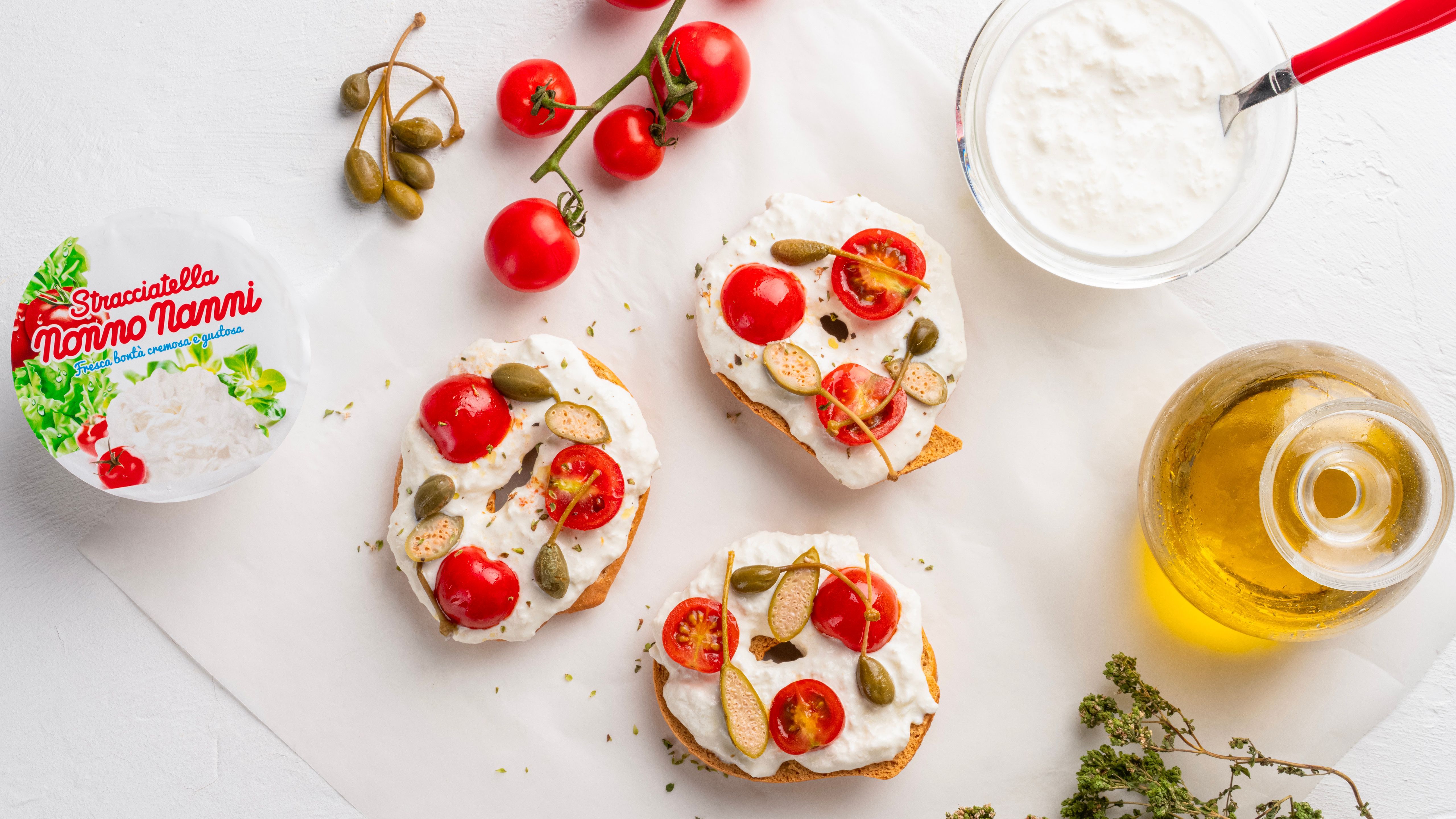 Crostini with stracciatella, cherry tomatoes, capers and oil next to a Nonno Nanni pack, on a white background.