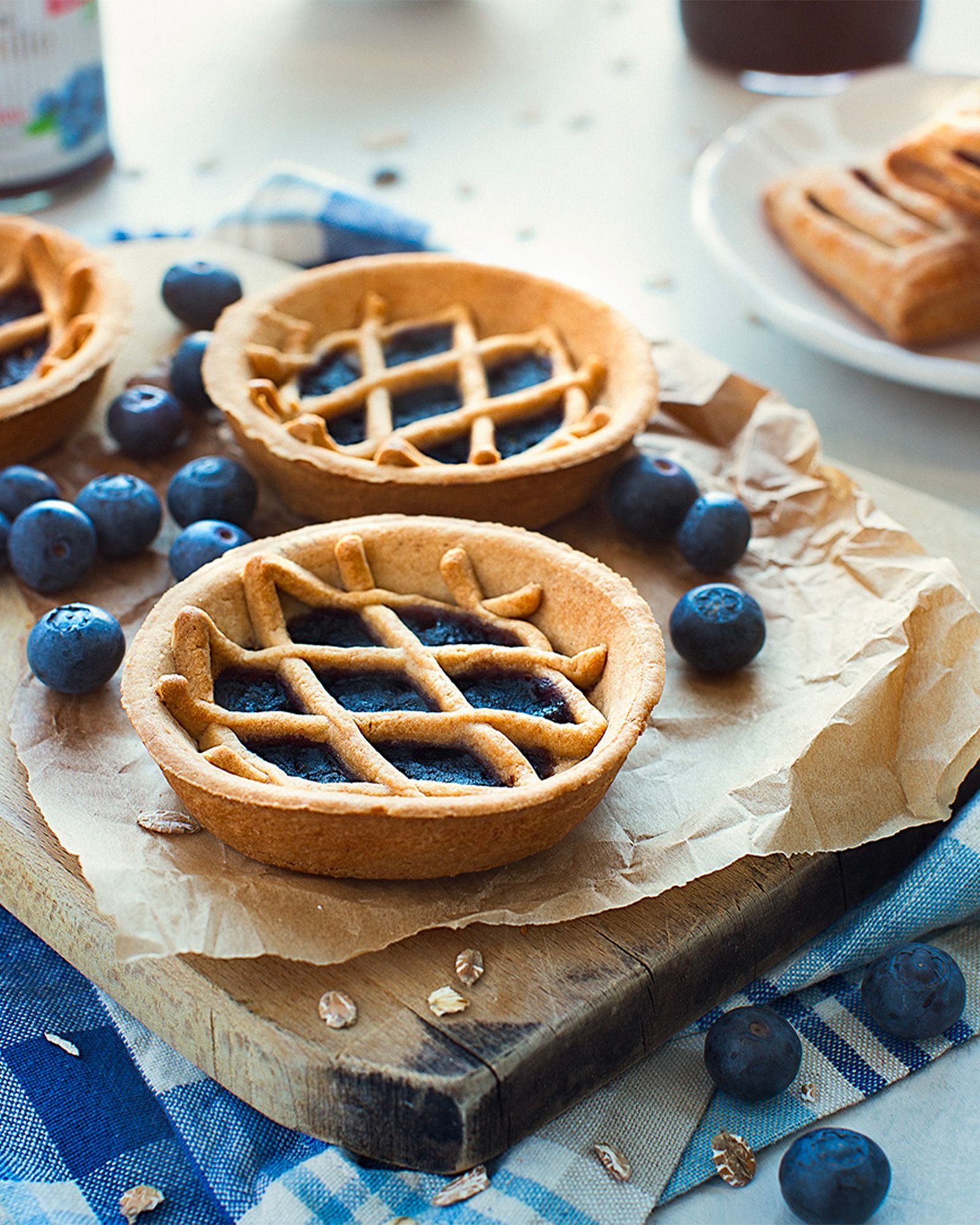 Blueberry tarts with a lattice shortcrust pastry top, surrounded by fresh blueberries on a wooden board with a blue checked tablecloth