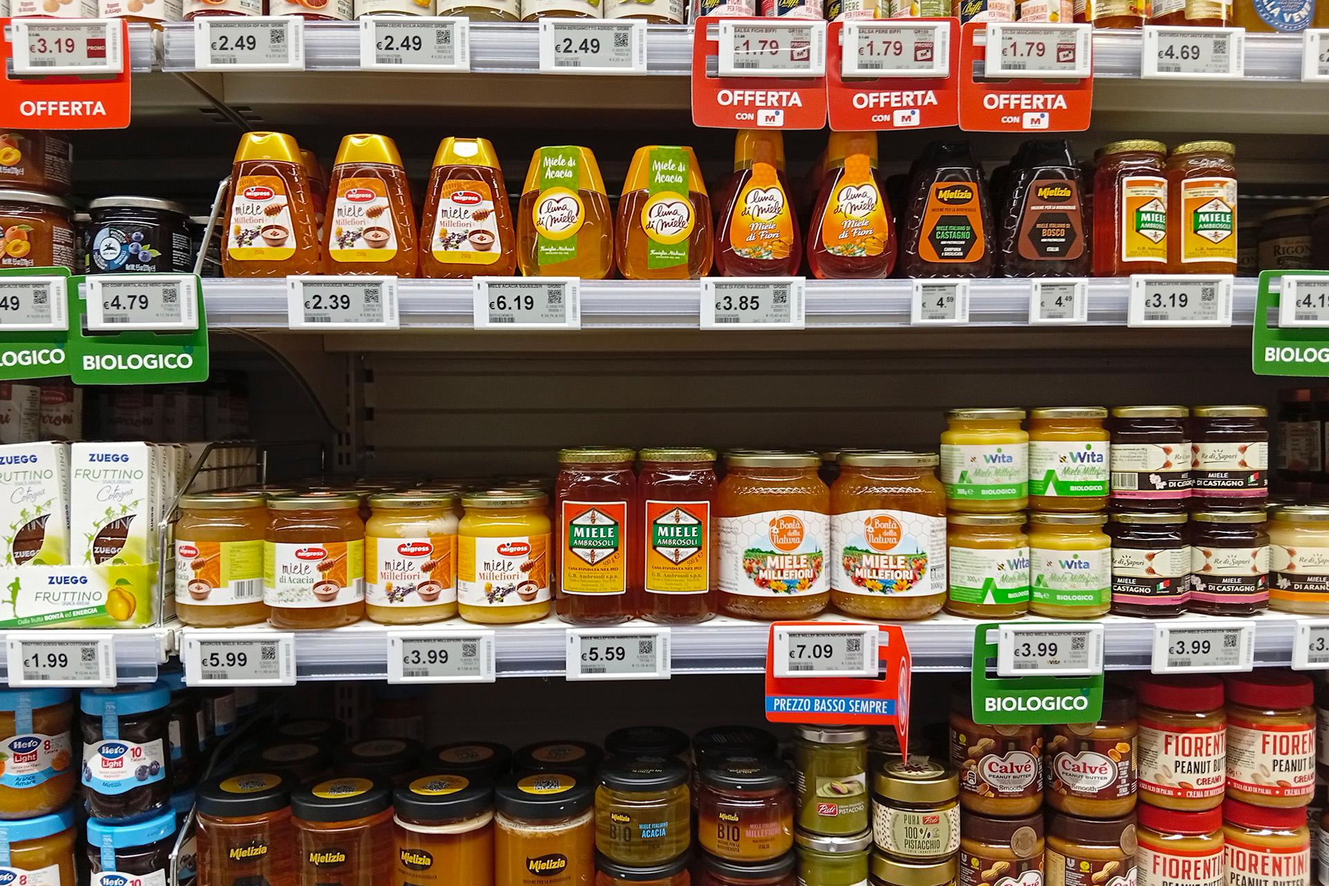 un-inquadratura-frontale-di-alcuni-scaffali-di-un-supermercato-ricchi-di-prodotti-alimentari-sul-primo-scaffale-in-alto-sono-esposti-vari-flaconi-di-miele-spremibili-tra-cui-miele-millefiori-e-miele-di-acacia-con-cartellini-rossi-delle-offerte-nello-scaffale-centrale-si-trovano-diversi-barattoli-di-vetro-di-miele-di-varie-marche-tra-cui-ambrosoli-e-wita-biologico-affiancati-a-sinistra-da-confezioni-di-fruttino-zuegg-lo-scaffale-inferiore-mostra-invece-altri-barattoli-di-miele-creme-spalmabili-come-quella-al-pistacchio-burro-d-arachidi-fiorentini-e-maionese-calvè