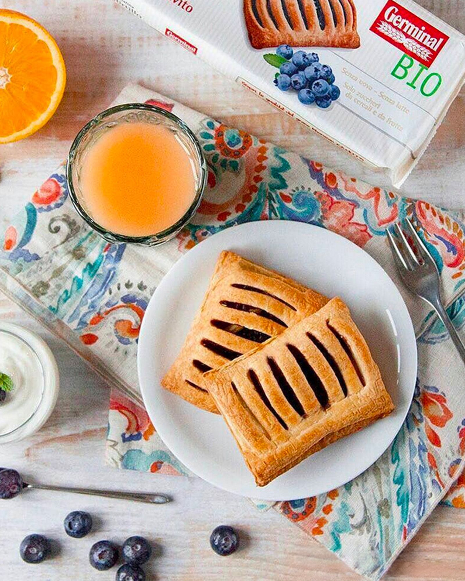 an overhead shot of a breakfast or snack scene with two filled puff-pastry parcels on a small white plate set on a floral-patterned cloth napkin, next to a glass of fresh orange juice, a small fork, half a fresh orange, a few scattered blueberries on the table, and a small bowl of yogurt, with the Germinal Bio product pack visible in the top right