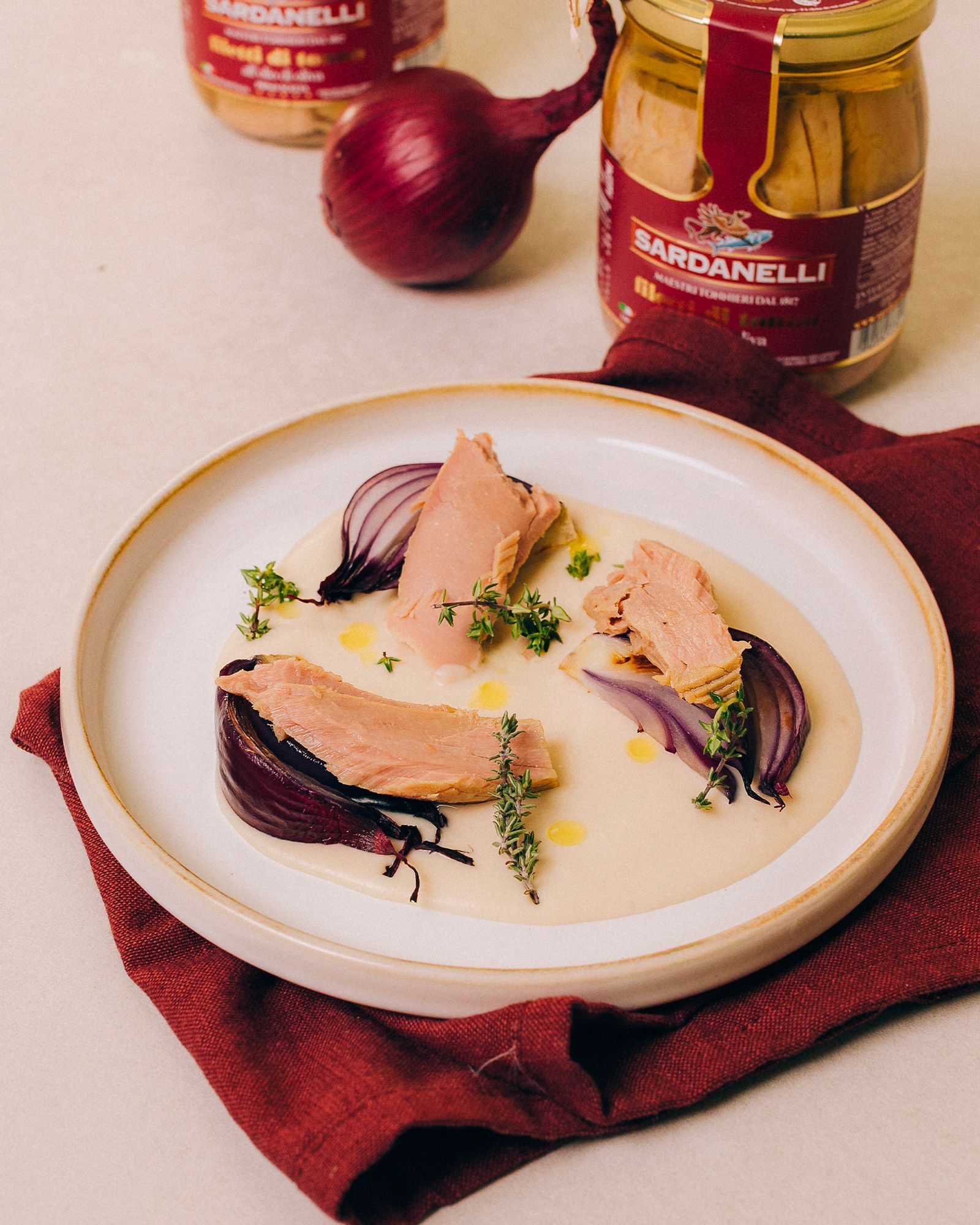 A plate with Sardanelli tuna fillets resting on a light cream, accompanied by petals of cooked red onion and thyme leaves. The plate sits on a burgundy cloth napkin, while a whole red onion and two glass jars of the product with the Sardanelli label appear blurred in the background.