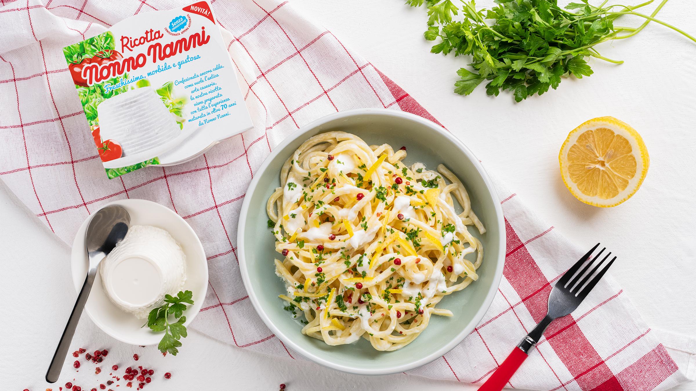 un-piatto-di-spaghetti-conditi-con-crema-di-ricotta-scorza-di-limone-pepe-rosa-e-prezzemolo-fresco-visto-dall-alto-su-un-tavolo-con-un-canovaccio-a-scacchi-rossi-e-bianchi-accanto-ci-sono-una-confezione-di-ricotta-nonno-nanni-un-piattino-con-una-ricottina-e-un-cucchiaio-mezzo-limone-una-forchetta-e-un-ciuffo-di-prezzemolo