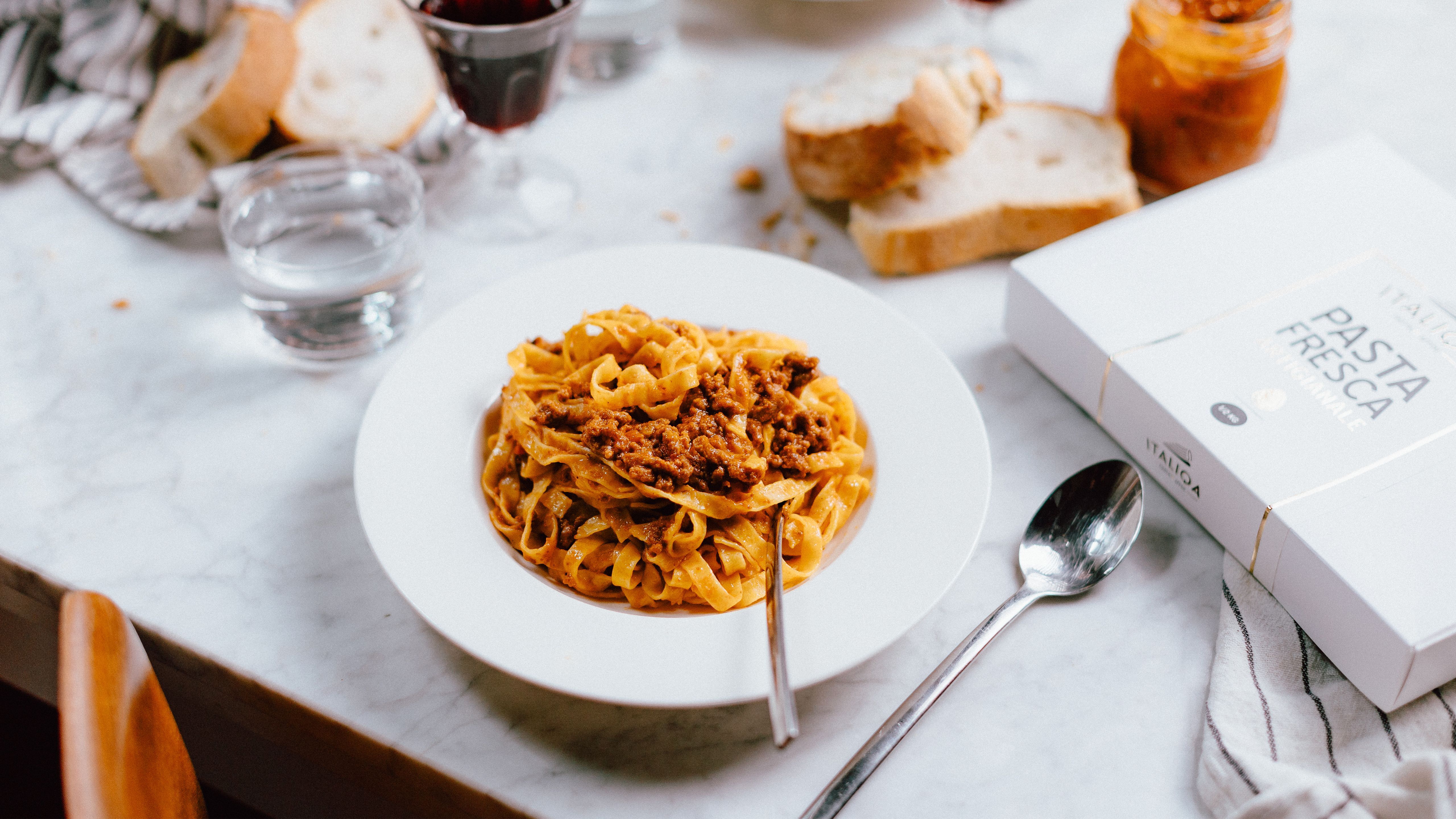 Tagliatelle with ragù on a white plate, next to a pack of Italiqa fresh pasta and bread on the table.