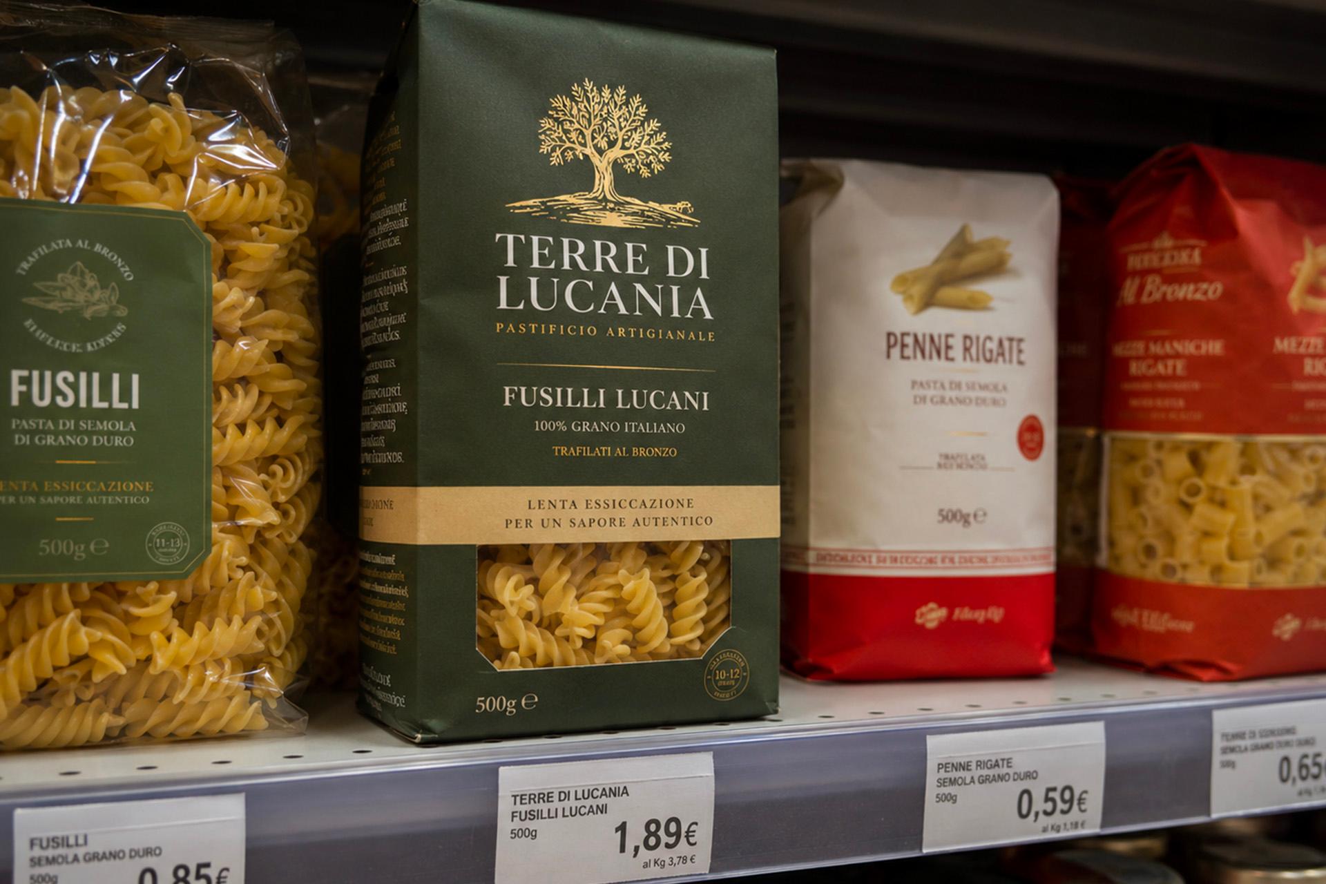 un-inquadratura-ravvicinata-di-uno-scaffale-di-un-supermercato-con-vari-prodotti-alimentari-in-alto-sono-esposte-diverse-confezioni-di-pasta-tra-cui-un-sacchetto-di-fusilli-trafilati-al-bronzo-da-ottantacinque-centesimi-una-scatola-verde-di-fusilli-lucani-marchiati-terre-di-lucania-pastificio-artigianale-al-prezzo-di-un-euro-e-ottantanove-e-un-sacchetto-di-penne-rigate-da-cinquantanove-centesimi-nella-parte-inferiore-dello-scaffale-sono-invece-allineati-diversi-barattoli-di-vetro-di-sugo-al-basilico