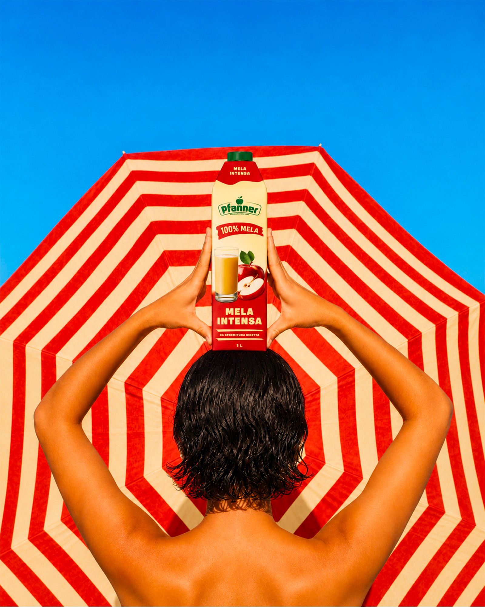 Person seen from behind holding a Pfanner Mela Intensa pack in front of a red-and-white striped beach umbrella against a blue sky