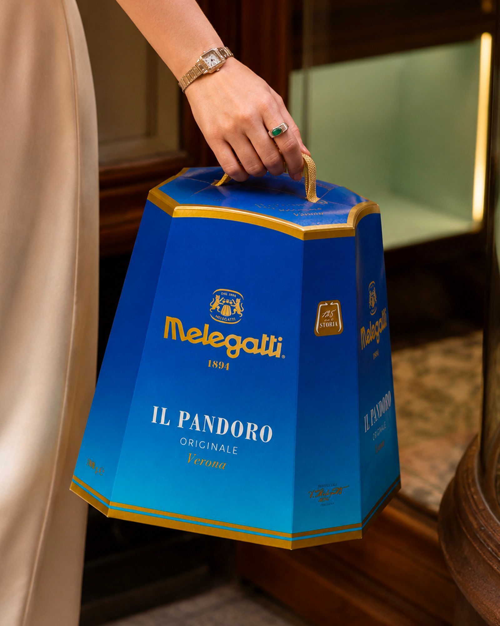 A person in light-colored clothing carrying a blue and gold Melegatti Pandoro Originale pack by its handle.