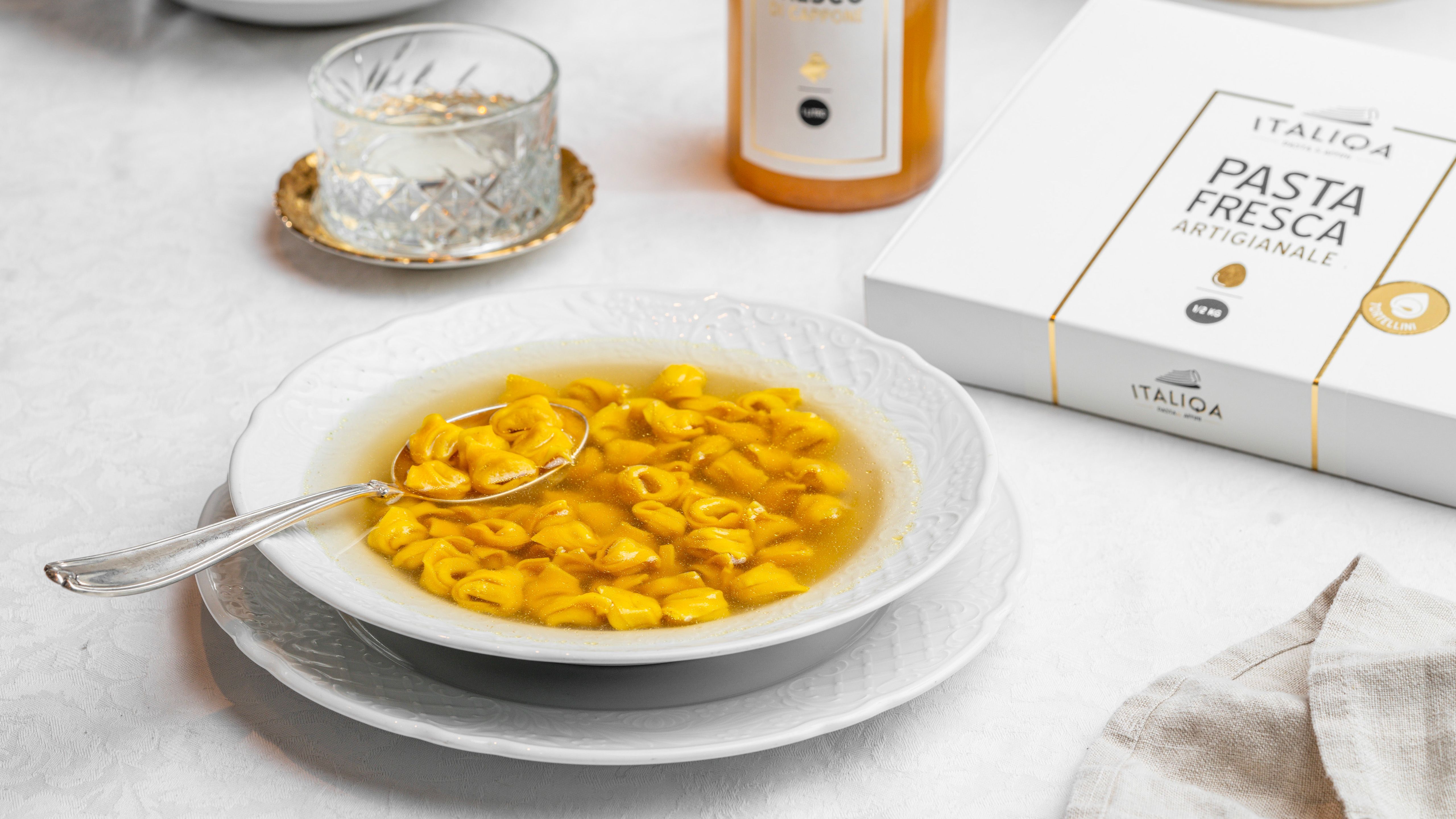 A plate of tortellini in broth next to a pack of Italiqa artisan fresh pasta, a glass and a bottle, on a white table.