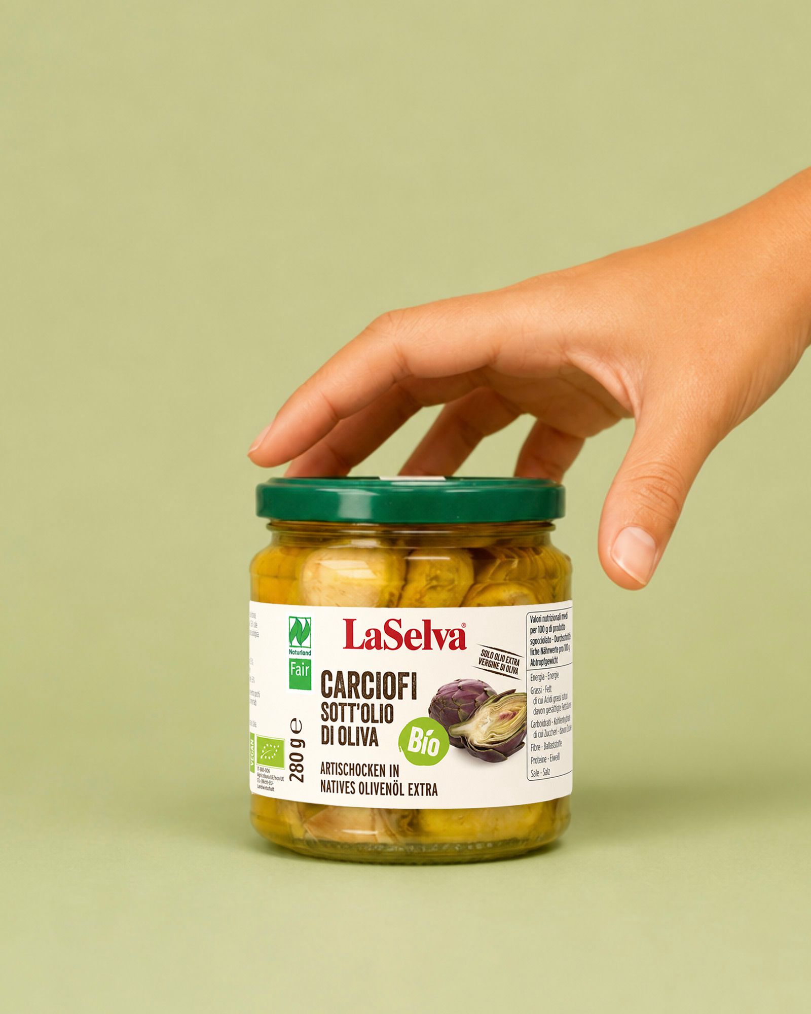 Jar of La Selva organic artichokes in oil with a hand on the lid, on a light green background