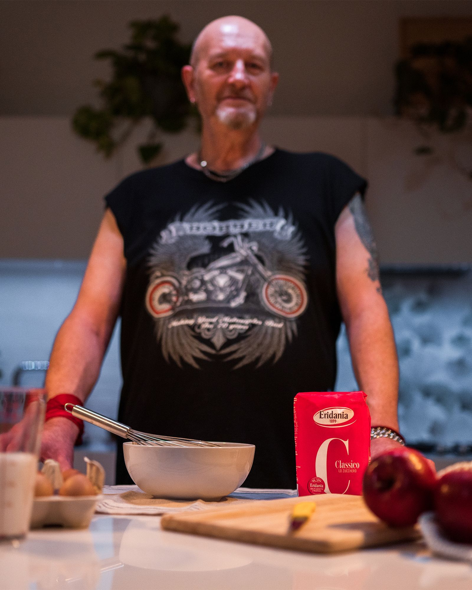 Man in a biker t-shirt behind a kitchen counter with Eridania Classico sugar, a whisk, eggs, milk and apples