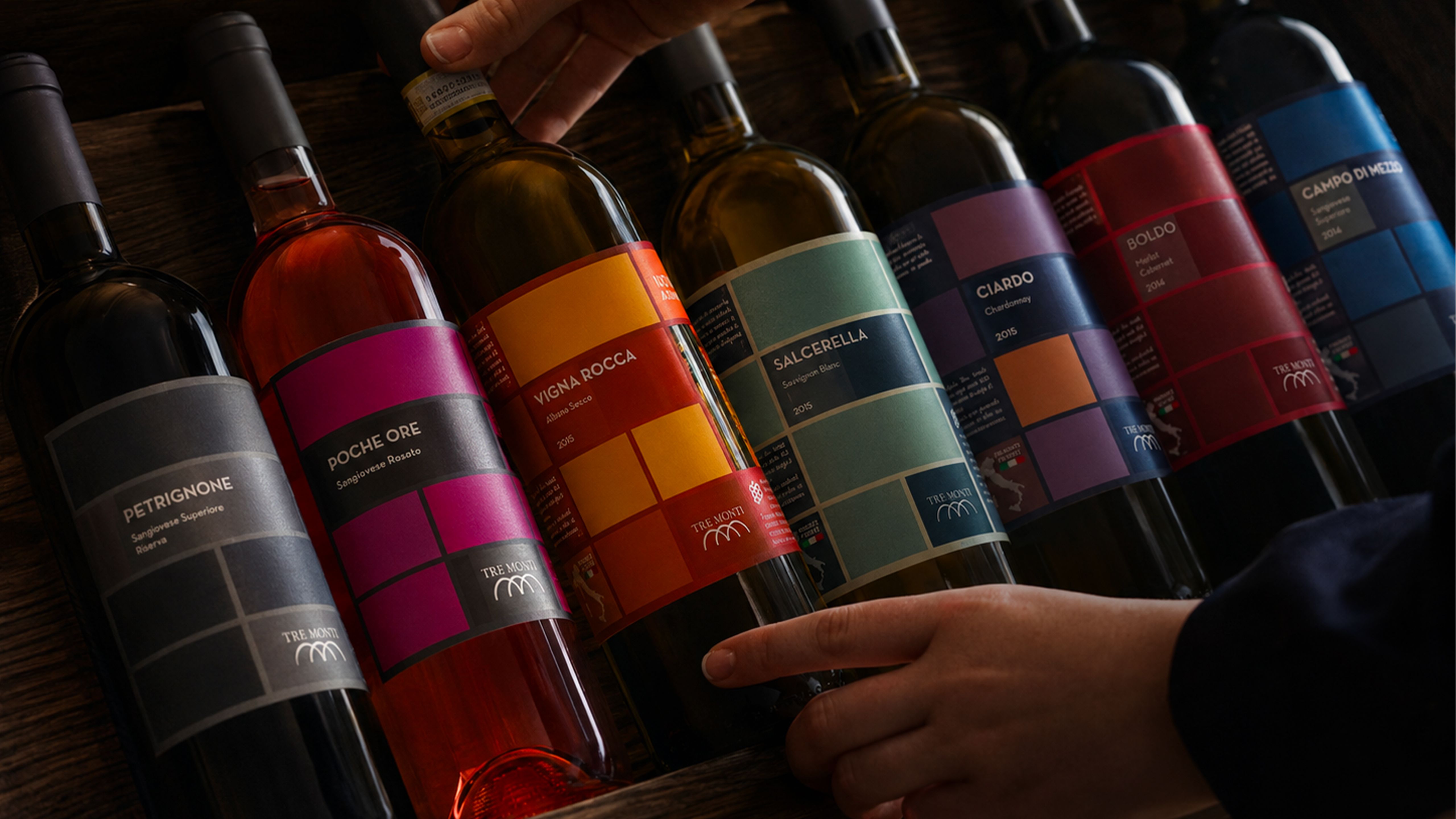 Bottles of Tre Monti wine with colorful labels arranged on a wooden surface, being selected by two hands.