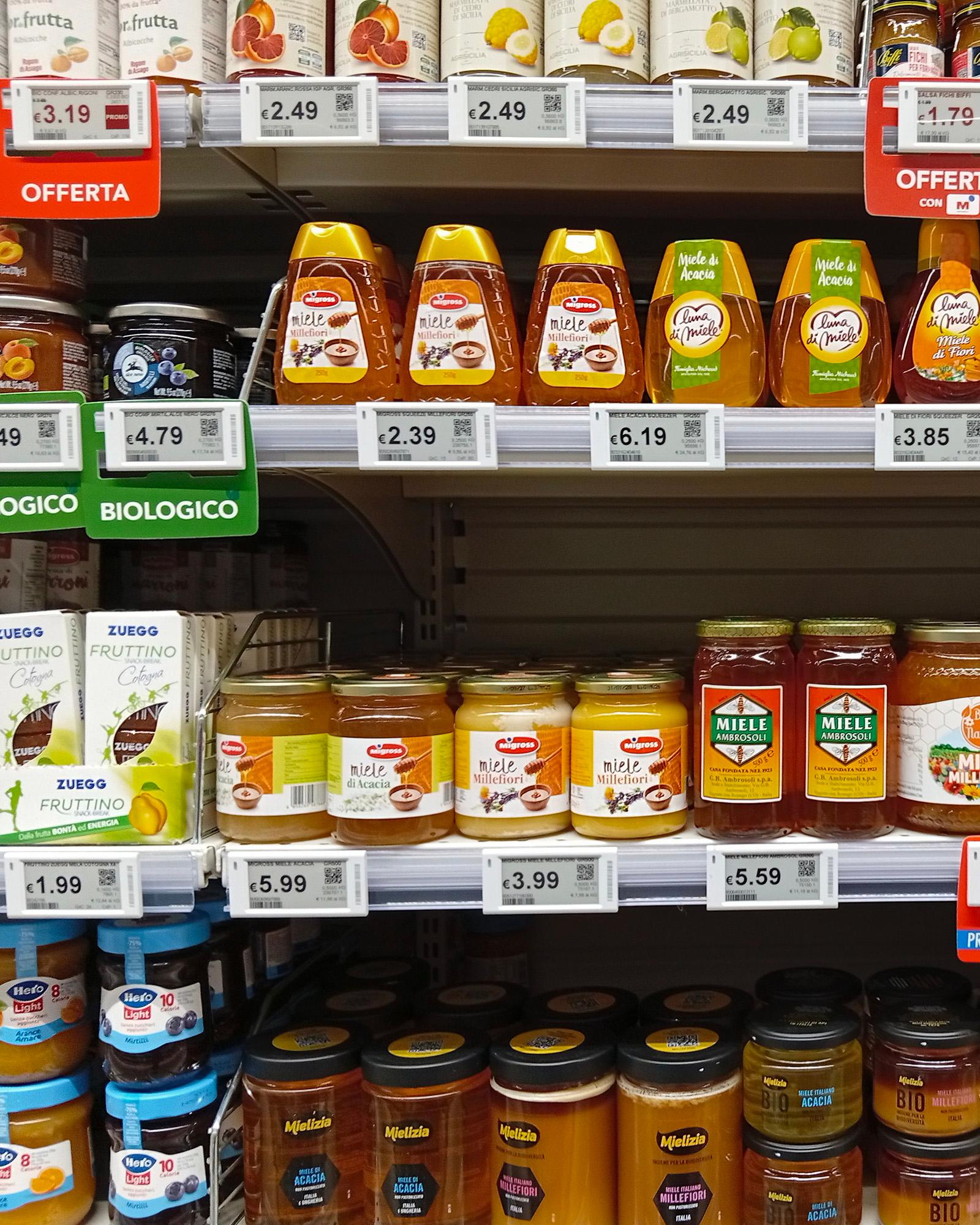 un-inquadratura-frontale-di-alcuni-scaffali-di-un-supermercato-ricchi-di-prodotti-alimentari-sul-primo-scaffale-in-alto-sono-esposti-vari-flaconi-di-miele-spremibili-tra-cui-miele-millefiori-e-miele-di-acacia-con-cartellini-rossi-delle-offerte-nello-scaffale-centrale-si-trovano-diversi-barattoli-di-vetro-di-miele-di-varie-marche-tra-cui-ambrosoli-e-wita-biologico-affiancati-a-sinistra-da-confezioni-di-fruttino-zuegg-lo-scaffale-inferiore-mostra-invece-altri-barattoli-di-miele-creme-spalmabili-come-quella-al-pistacchio-burro-d-arachidi-fiorentini-e-maionese-calvè