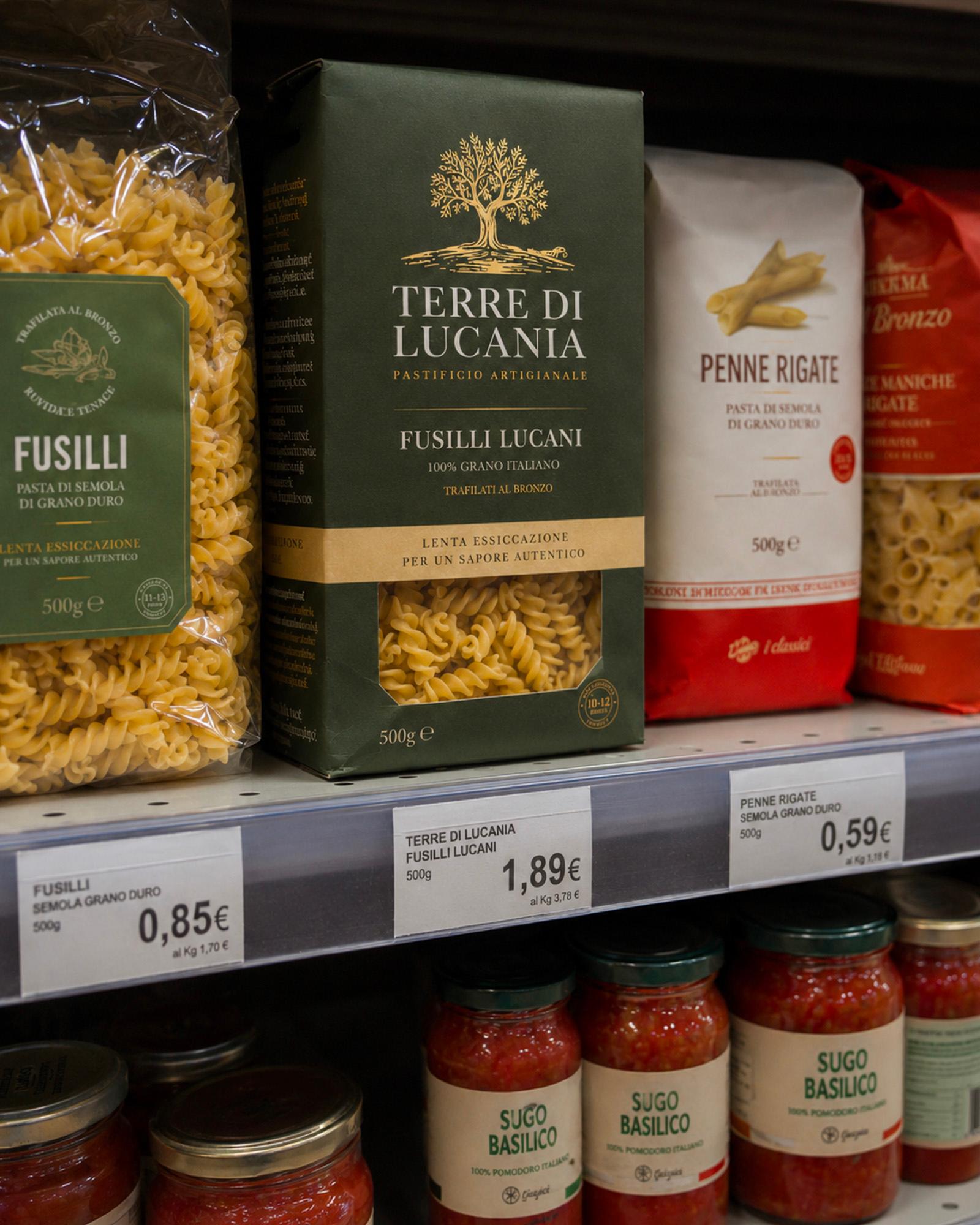 un-inquadratura-ravvicinata-di-uno-scaffale-di-un-supermercato-con-vari-prodotti-alimentari-in-alto-sono-esposte-diverse-confezioni-di-pasta-tra-cui-un-sacchetto-di-fusilli-trafilati-al-bronzo-da-ottantacinque-centesimi-una-scatola-verde-di-fusilli-lucani-marchiati-terre-di-lucania-pastificio-artigianale-al-prezzo-di-un-euro-e-ottantanove-e-un-sacchetto-di-penne-rigate-da-cinquantanove-centesimi-nella-parte-inferiore-dello-scaffale-sono-invece-allineati-diversi-barattoli-di-vetro-di-sugo-al-basilico
