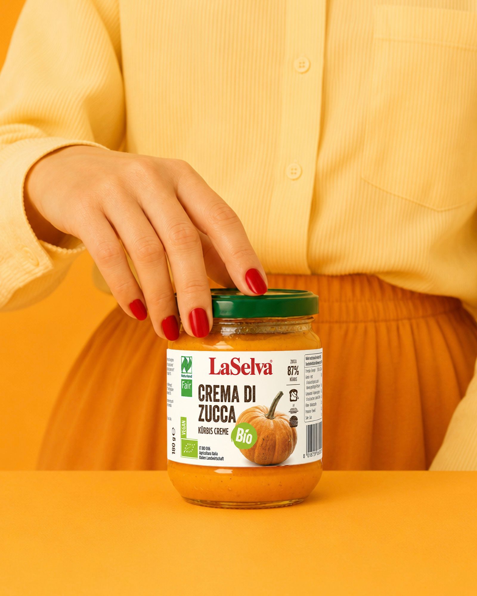 Person in a yellow shirt and orange skirt presenting a jar of La Selva organic pumpkin cream on an orange background