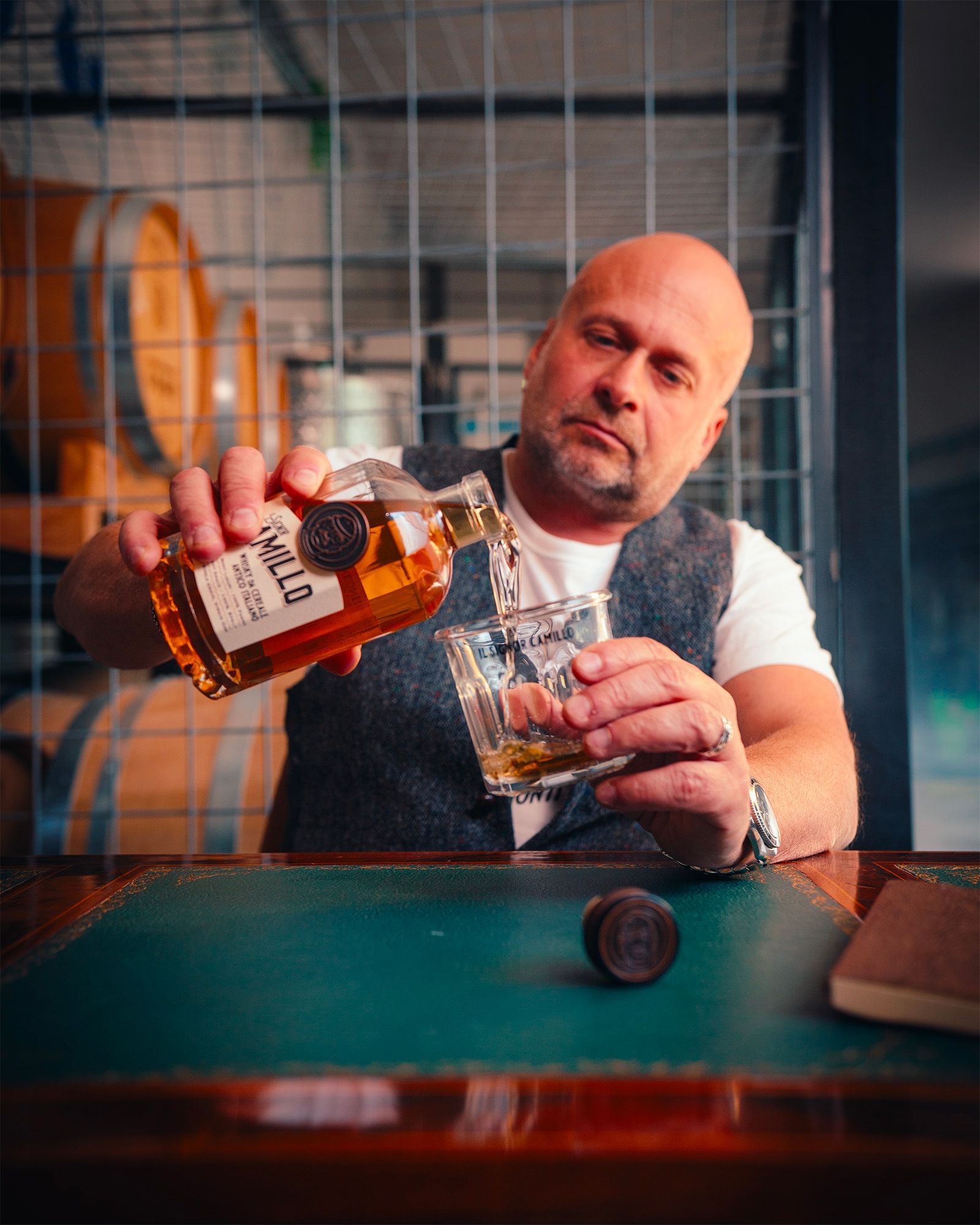 Man pouring a spirit from an Il Signor Camillo bottle into a glass, with wooden barrels in the background