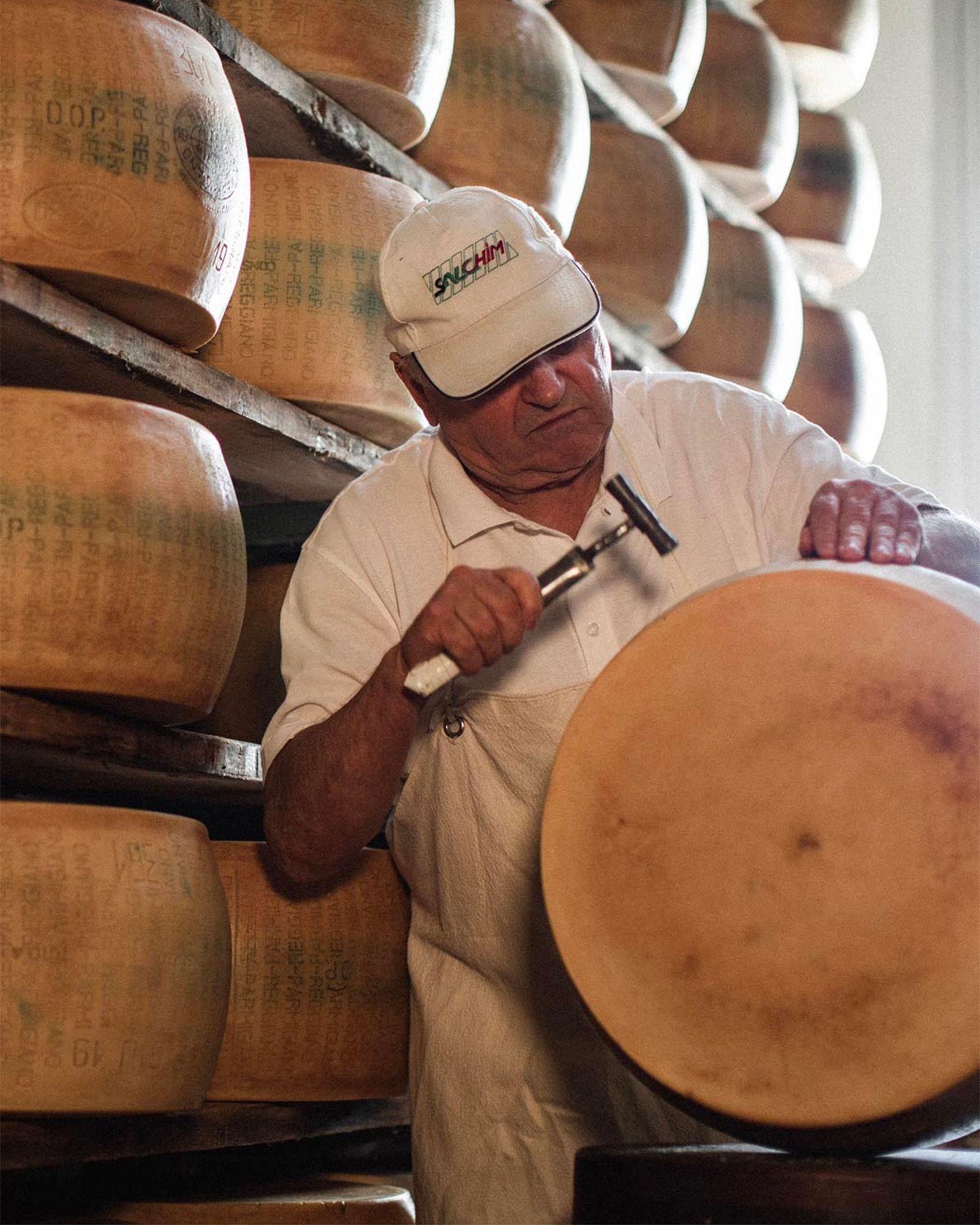 Cheesemaker tapping a wheel of Parmigiano Reggiano with a small hammer in the aging warehouse