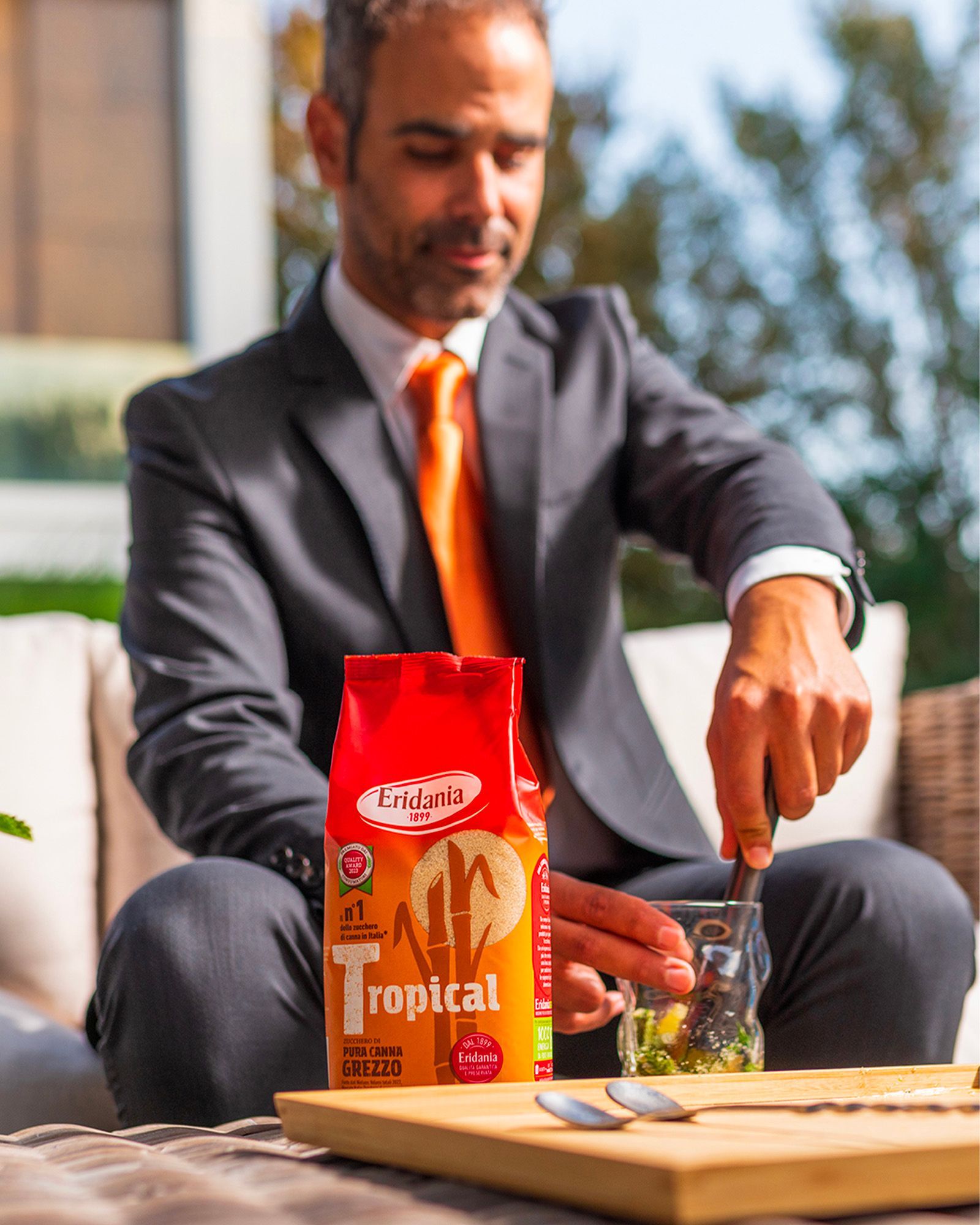 Man in a dark suit and orange tie stirring a drink in a glass next to a pack of Eridania Tropical raw cane sugar on an outdoor table