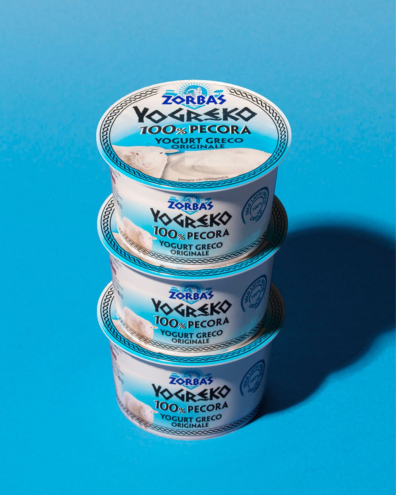 Three packs of Zorbas Yogreko 100% sheep's milk Greek yogurt stacked vertically on a clean light-blue background with a bold shadow on the right. The white and blue packaging shows the Zorbas logo, the wording 'Yogreko 100% Pecora – Original Greek Yogurt', and an illustration of a sheep.