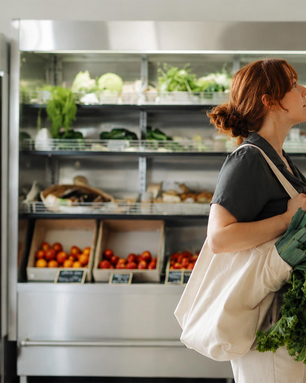 donna che osserva prodotti sugli scaffali di un negozio di alimentari con borsa in tela riutilizzabile contenente porri e cavolo riccio davanti a un banco refrigerato con frutta e verdura fresca sullo sfondo