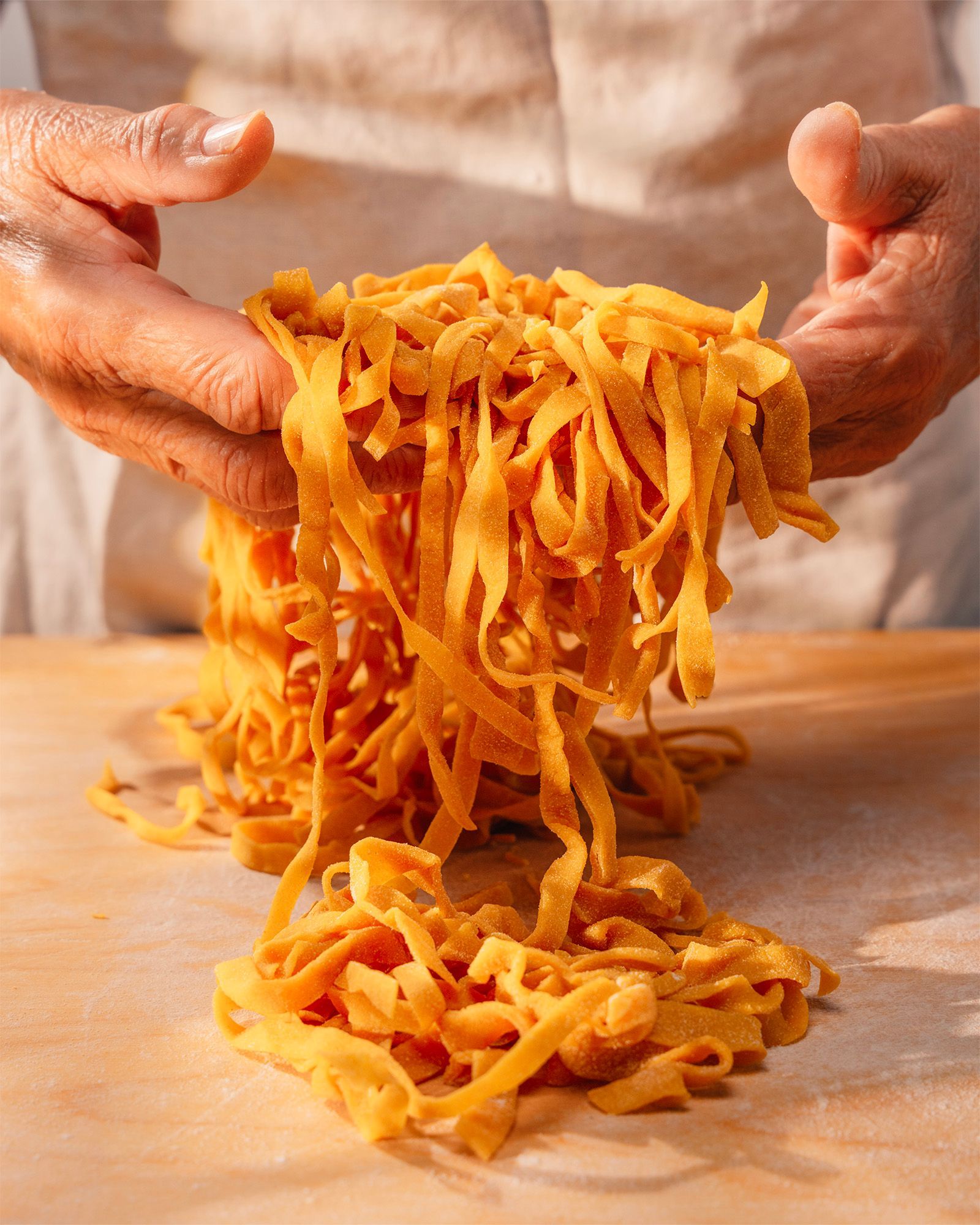 Elderly hands lifting homemade fresh egg pasta, tagliatelle-style, from a wooden pastry board
