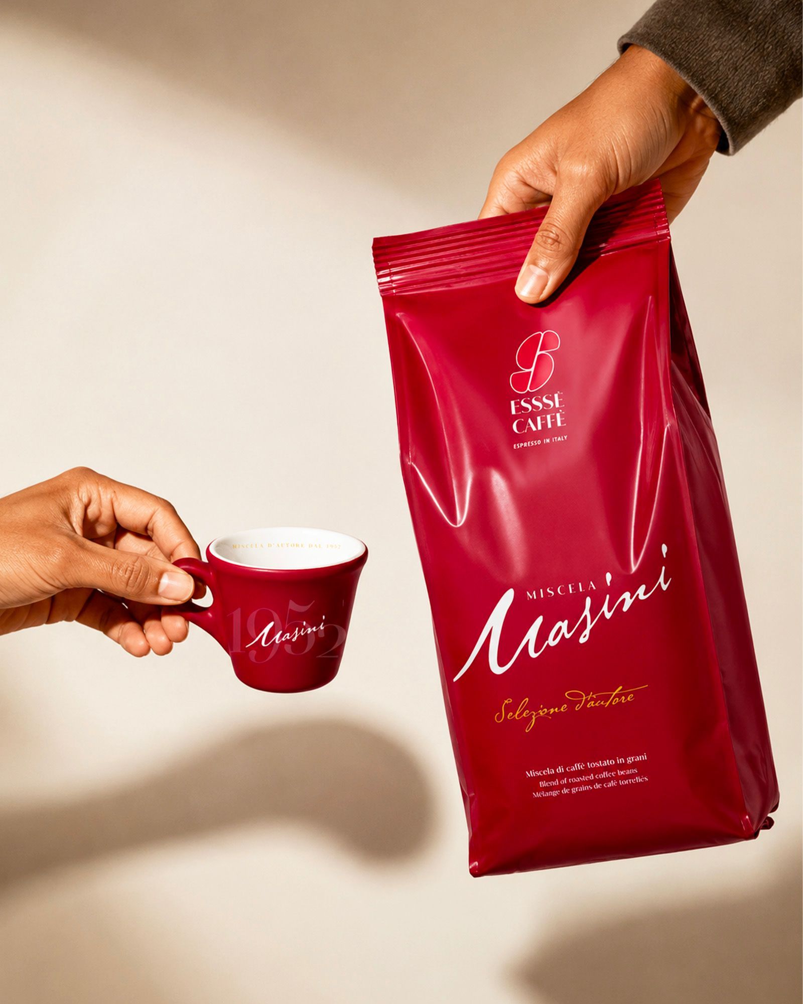 Two hands framed against a light background — one holding a red espresso cup with the Masini logo, the other holding a matching bag of whole bean coffee in the same color, from Essse Caffè's Masini blend line, labeled Selezione d'Autore.
