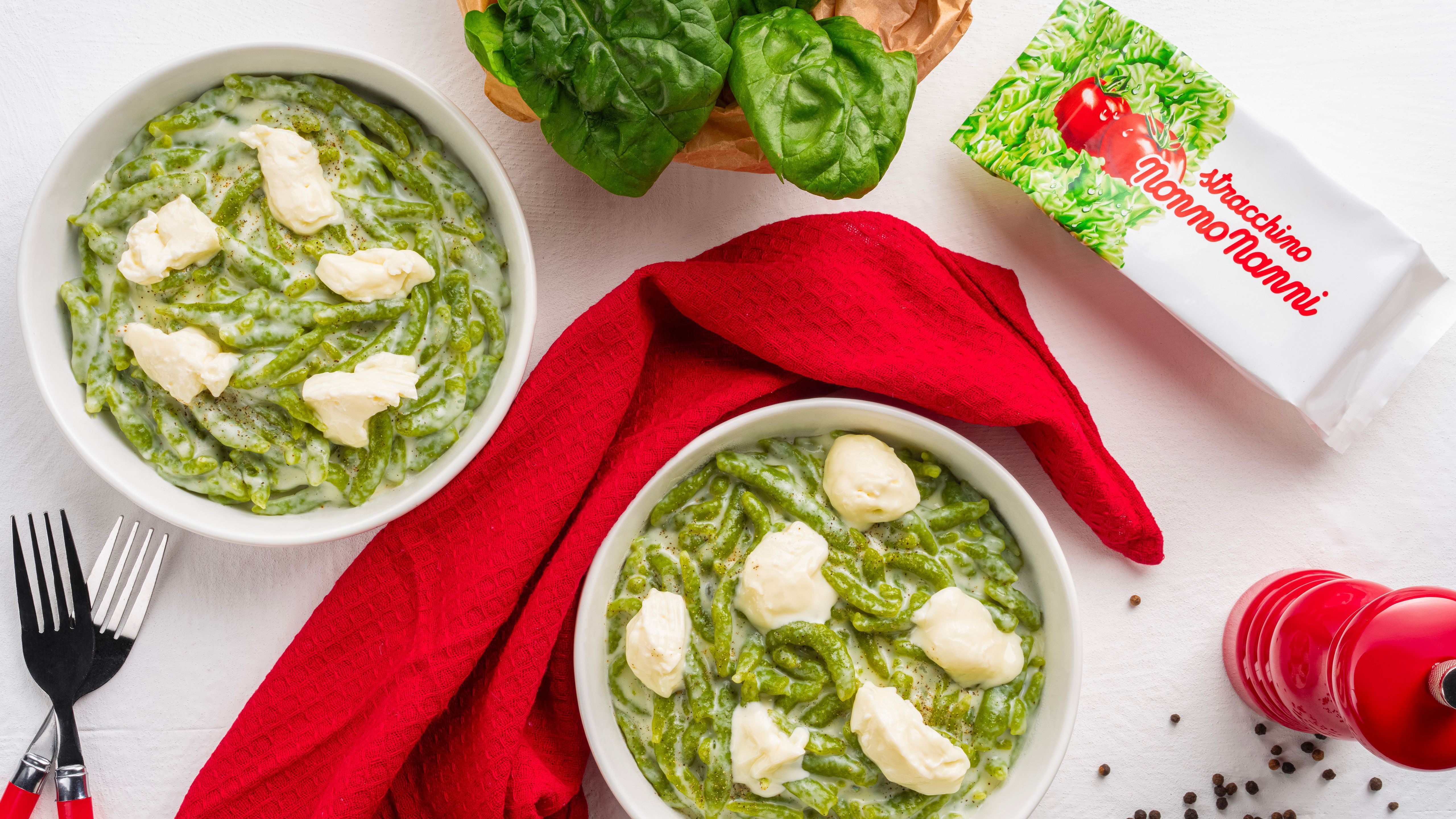 Two bowls of green pasta with cream and Nonno Nanni stracchino, next to basil, a red napkin, cutlery and a pepper mill, on a white background.