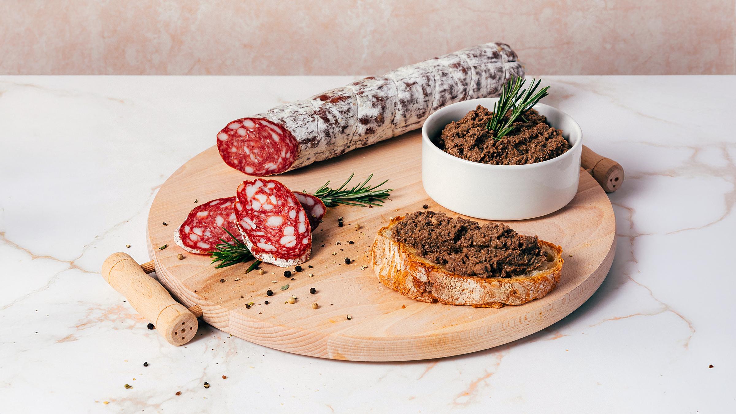 tagliere-di-legno-rotondo-con-salame-intero-affettato-crostino-di-pane-con-patè-e-ciotola-di-patè-con-rosmarino-su-tavolo-di-marmo