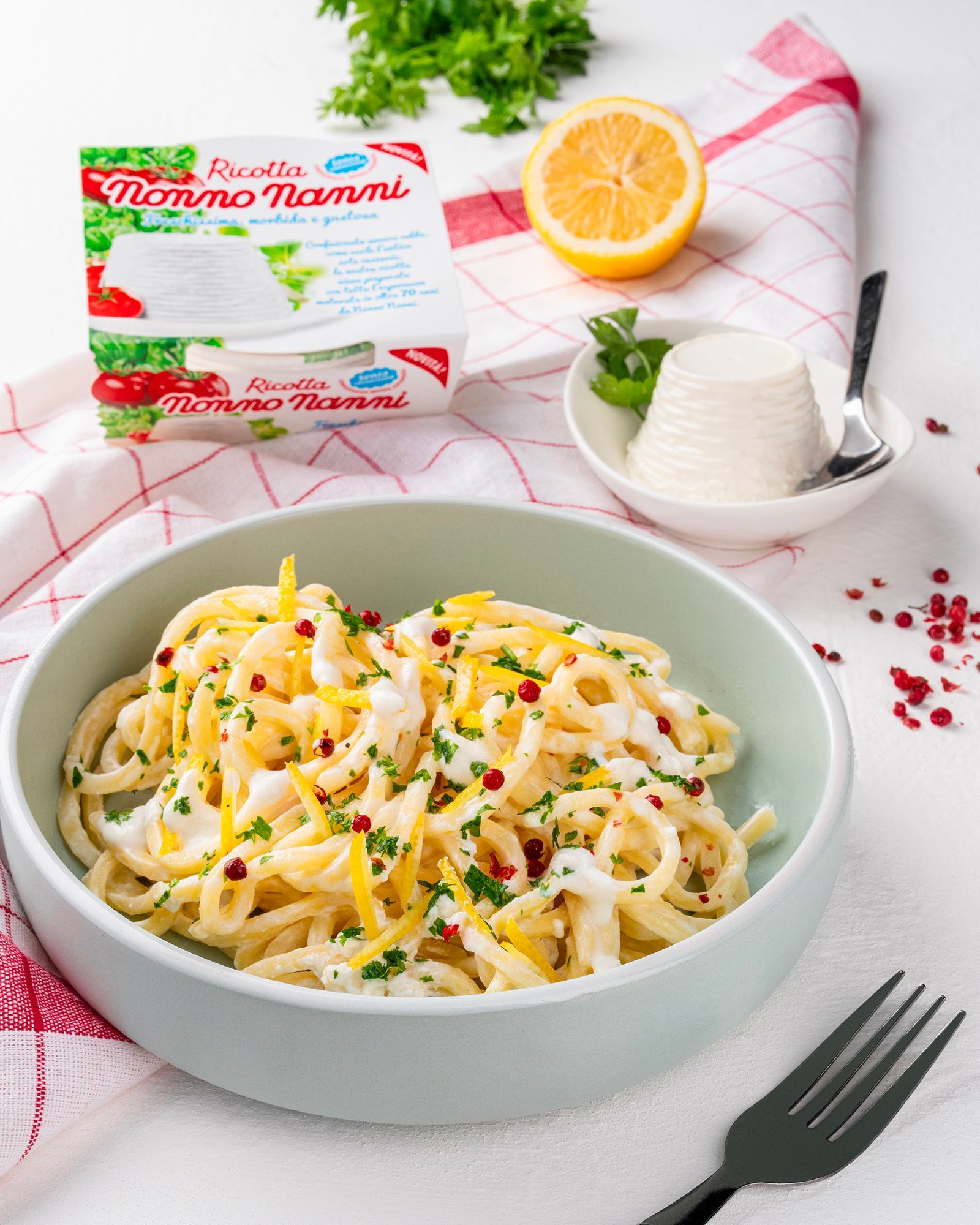 Creamy spaghetti with ricotta, lemon, parsley and pink pepper next to a pack of Nonno Nanni ricotta, on a white table.