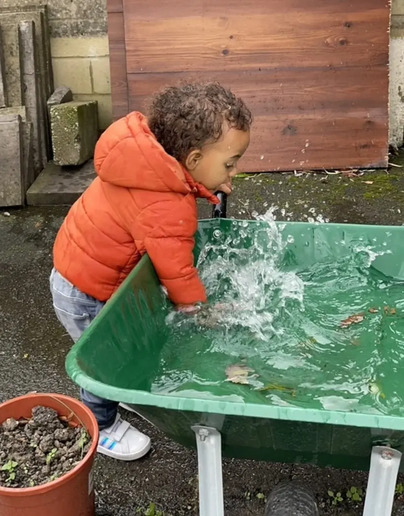 child-in-orange-jacket-playing-with-water-in-green-wheelbarrow