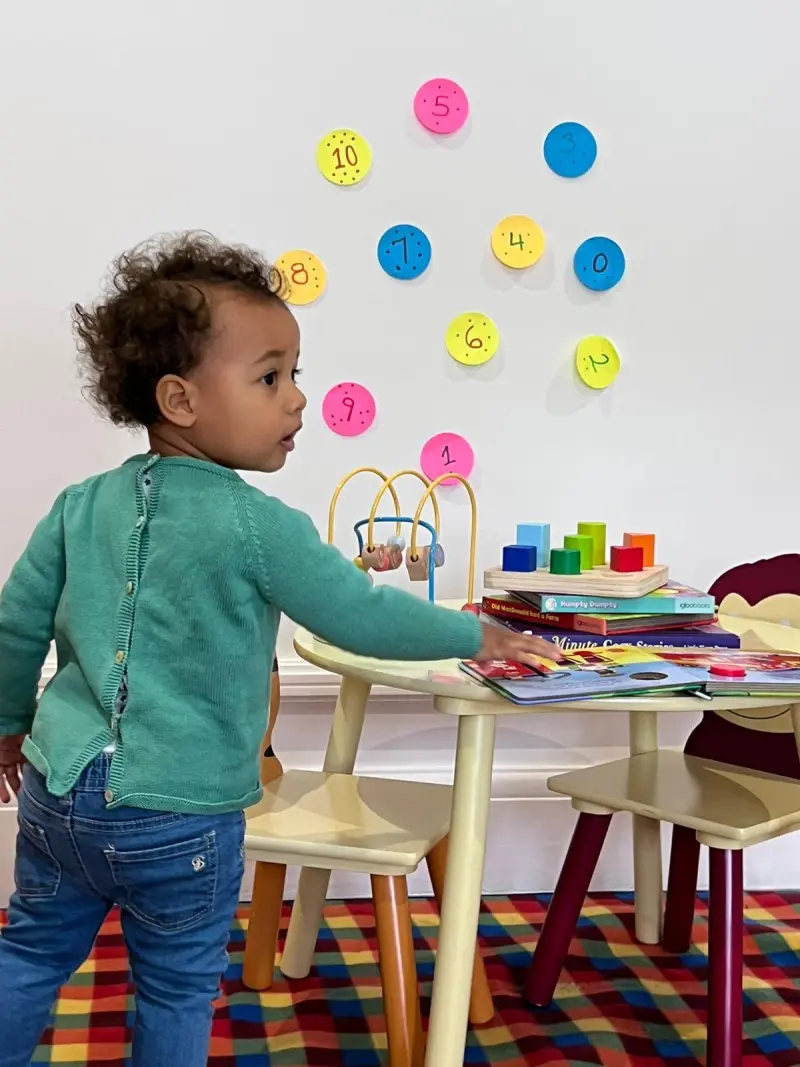 toddler-explores-colorful-toys-at-small-table-numbers-on-wall-behind