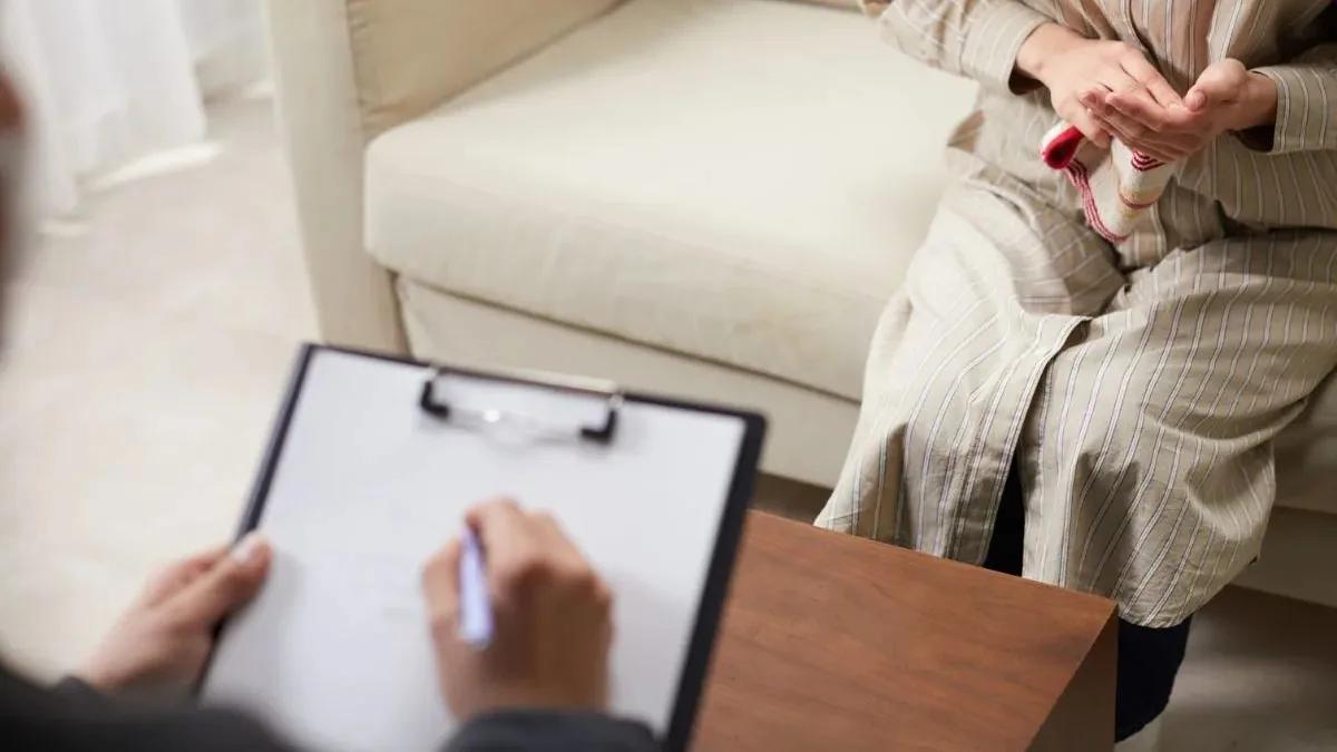 A clinician writing notes on a clipboard while a client sits on a sofa opposite
