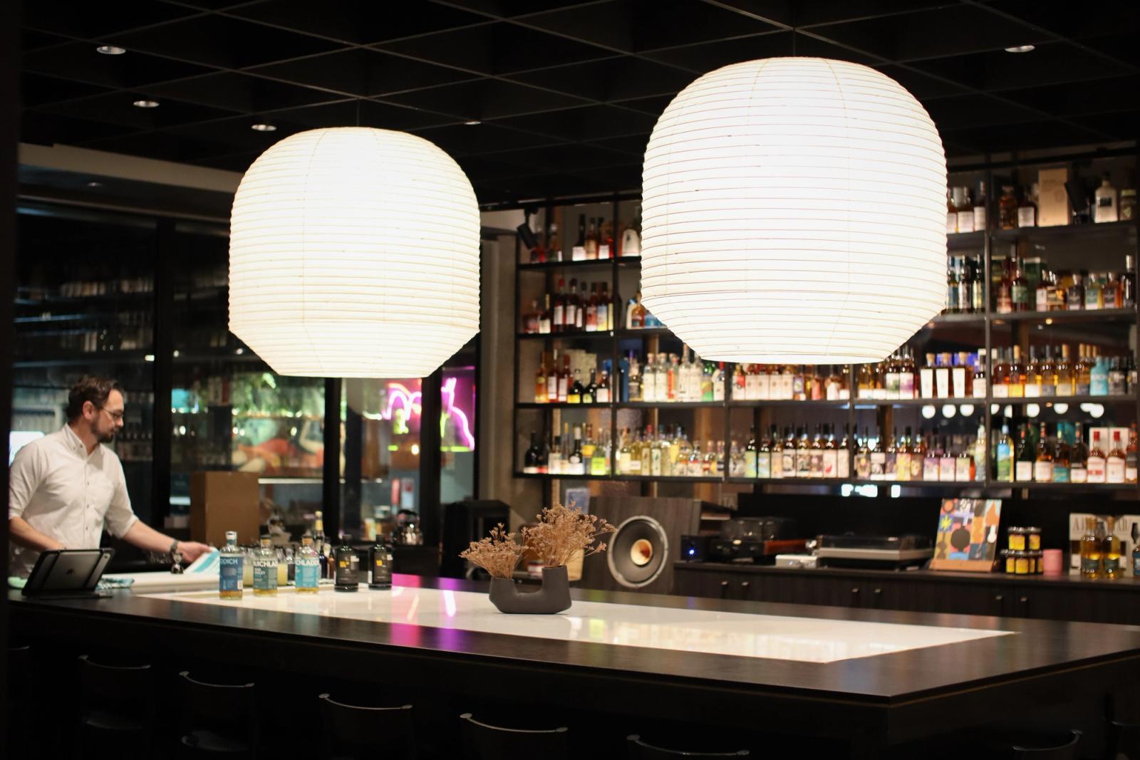 barman praparing cocktails pendants susppended over an island bar within tasting room display shelving in the background